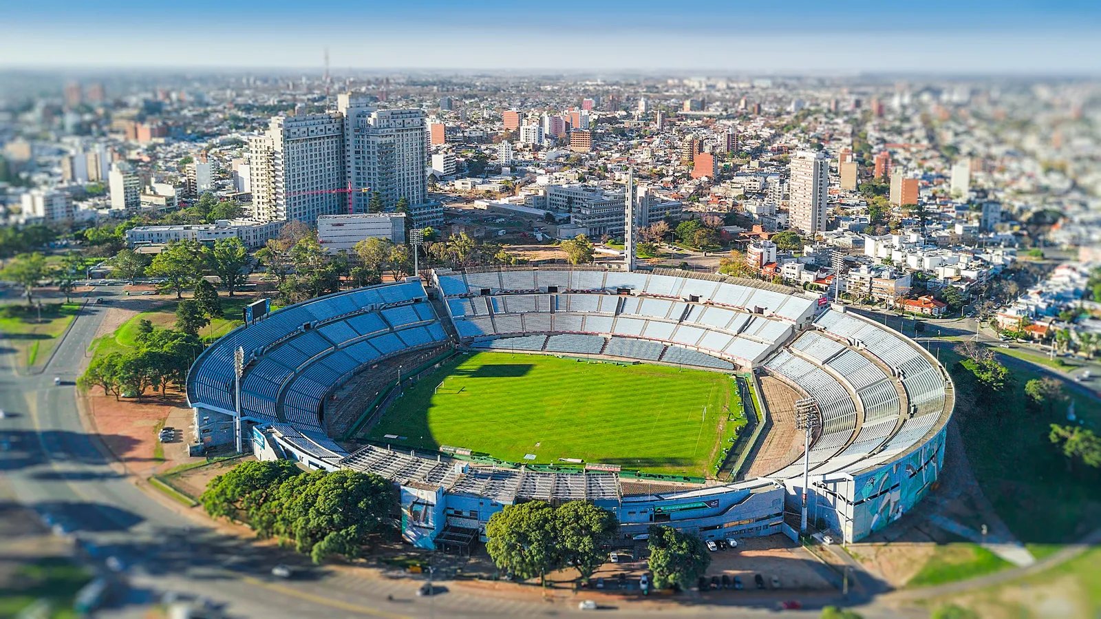 An aerial view of the Estadio Centenario in Montevideo, its oval bowl of pale terraces around a green pitch, ringed by the trees of Parque Batlle with the city's apartment blocks behind