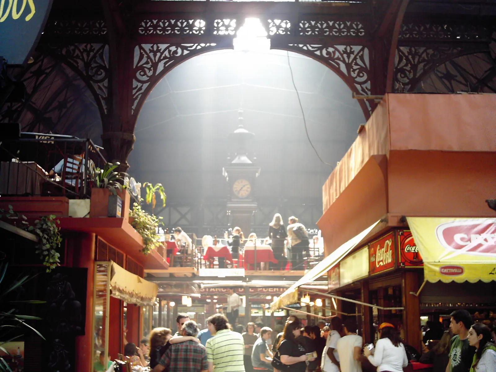 Inside the Mercado del Puerto in Montevideo, looking down the hall under a dark wrought-iron arch towards the market's clock, with grill smoke hanging in the light and people eating at tables on both sides