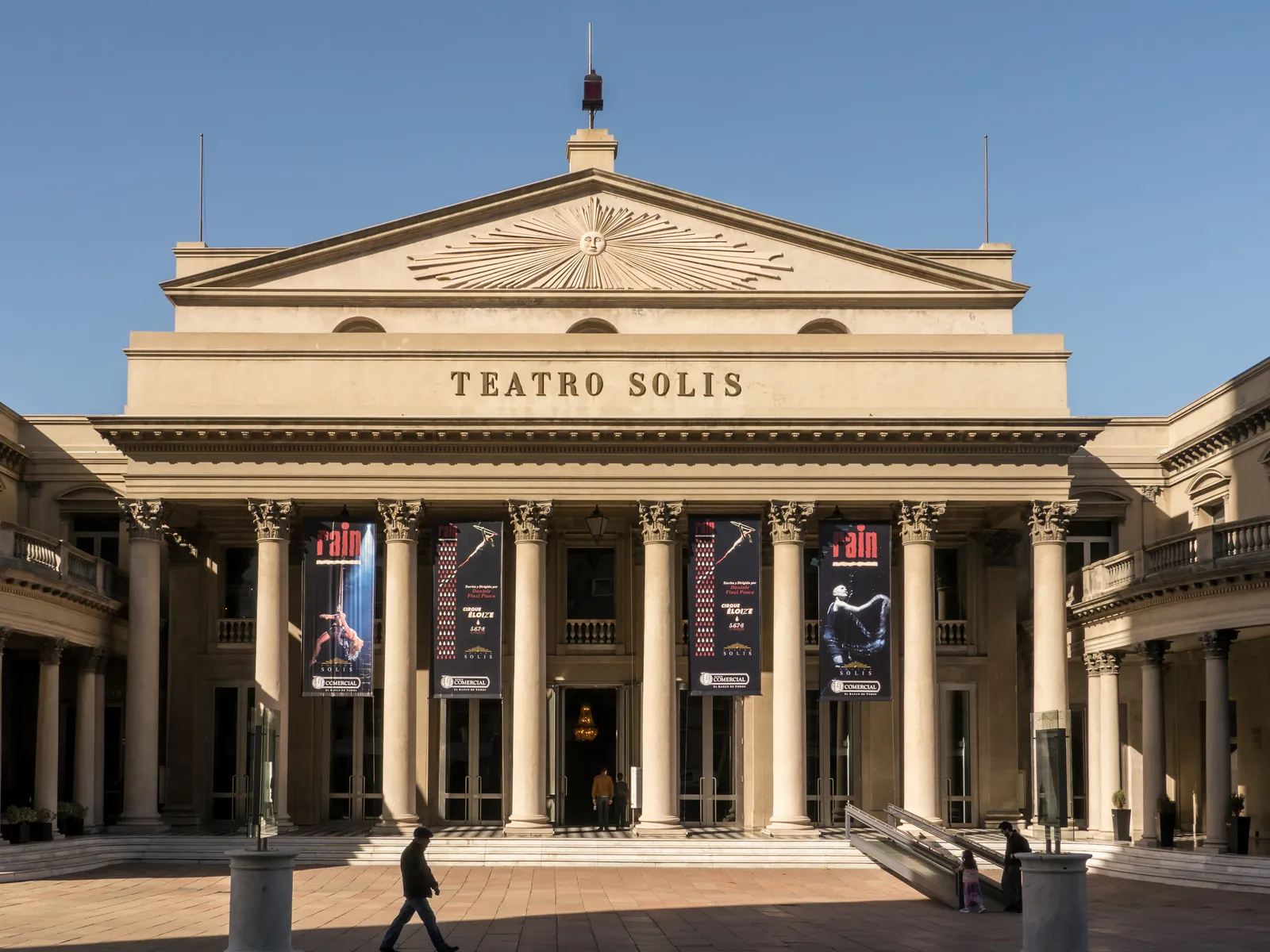 The colonnaded front of Teatro Solís in Montevideo, its name carved across the entablature above eight tall columns, with theatre banners hanging between them and a paved square in front