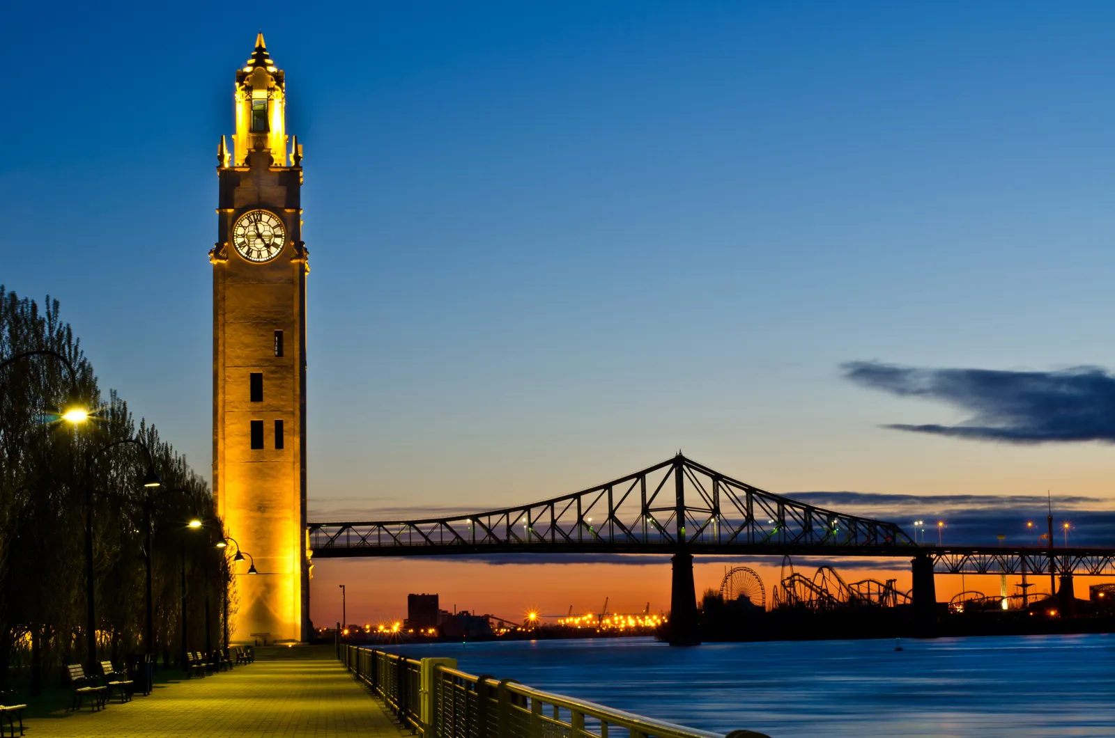 The lit clock tower at the eastern end of Montreal's Old Port quays at first light, with the steel span of the Jacques Cartier Bridge crossing the St Lawrence behind it