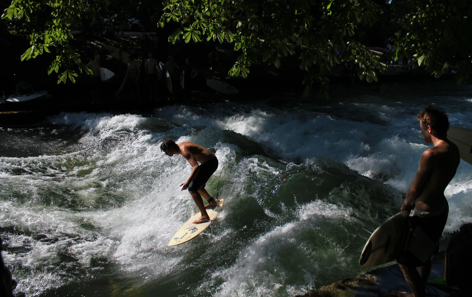 A surfer riding the standing wave on the Eisbach in Munich's Englischer Garten, another surfer waiting at the bank with a board, trees overhanging the water