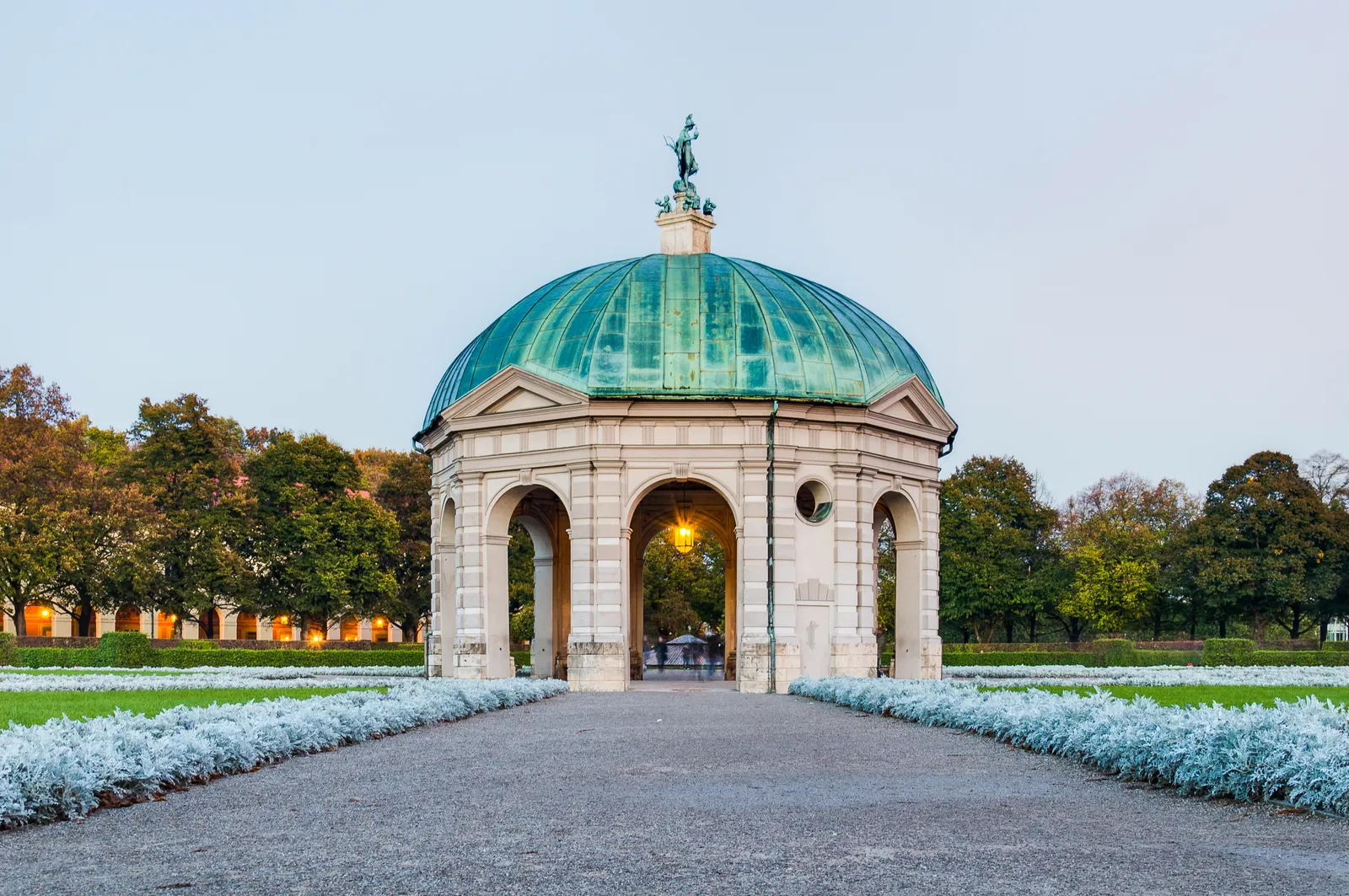 The Dianatempel pavilion at the centre of Munich's Hofgarten, an open-arched stone rotunda under a green copper dome, with gravel paths and low planting running toward it