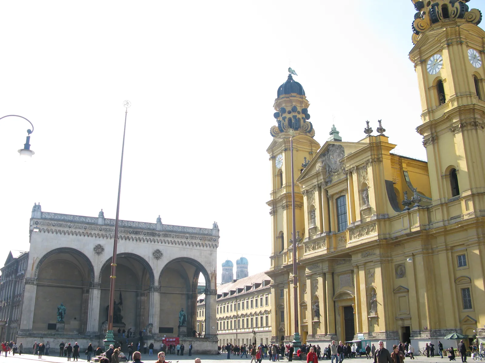 Odeonsplatz in Munich in daylight, the arched Feldherrnhalle on the left and the yellow Baroque towers of the Theatinerkirche on the right, with people crossing the open square between them