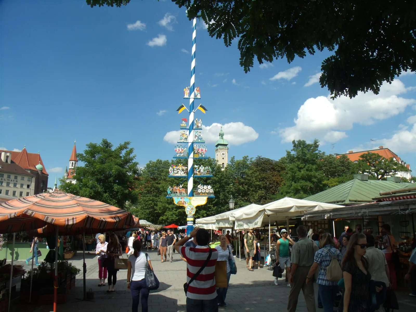 The Viktualienmarkt in Munich with its blue-and-white striped maypole rising above the stalls and sun umbrellas, shoppers moving between them and the tower of the Altes Rathaus behind