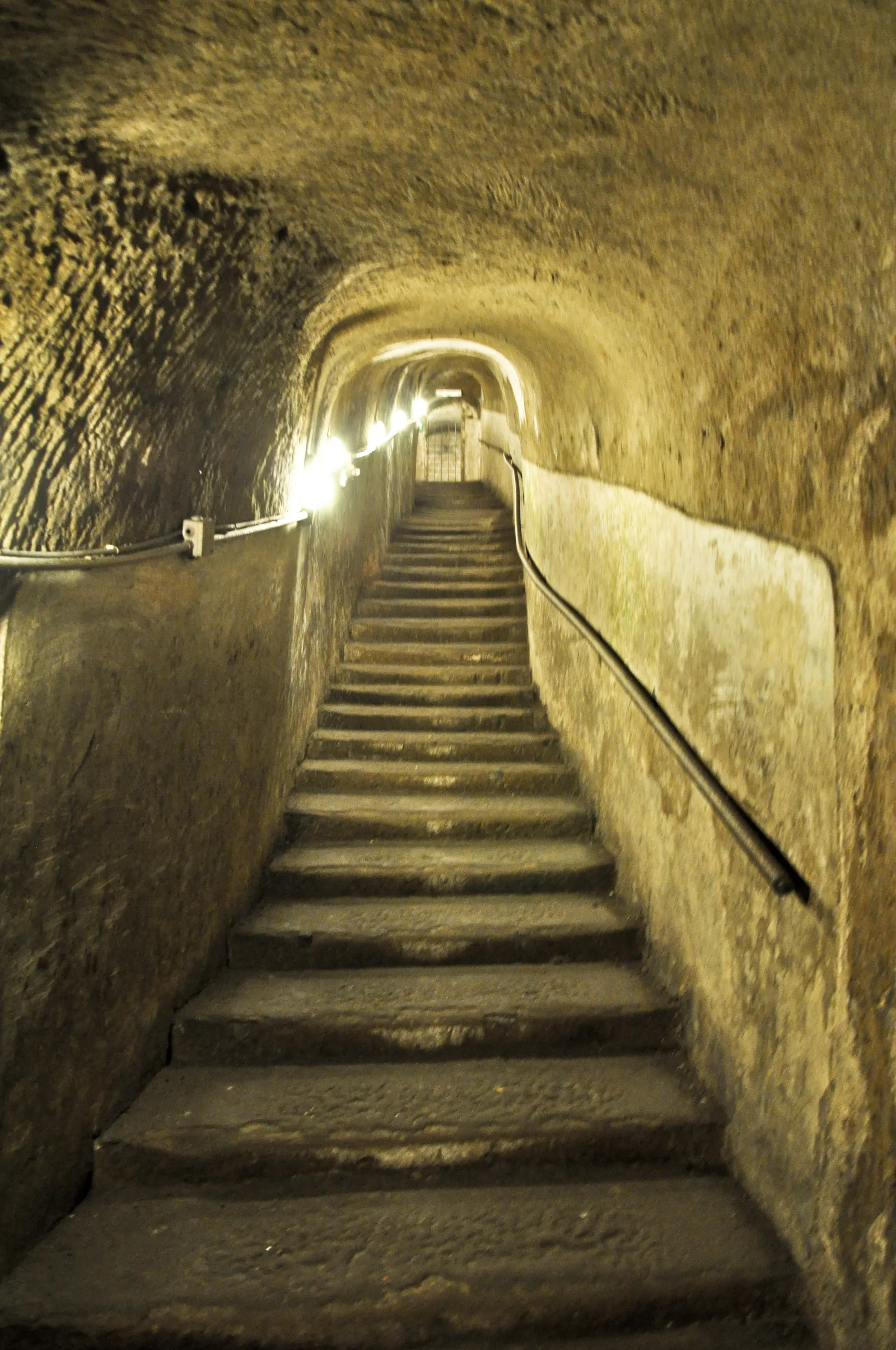 A stone stairway climbing through a lit tunnel cut into the tuff under Naples