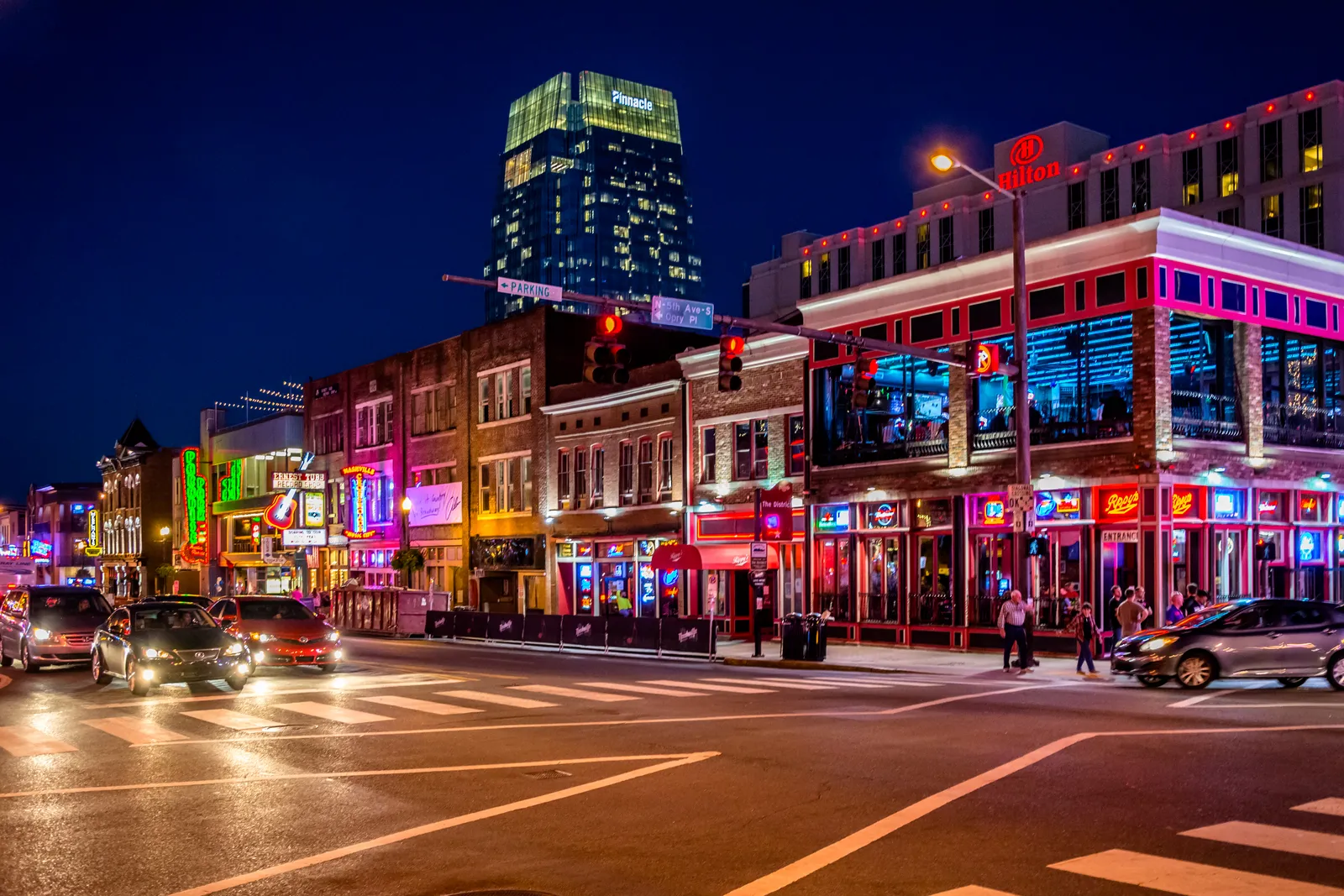 Lower Broadway in Nashville after dark, both sides of the street stacked with lit neon bar signs and an office tower rising behind them