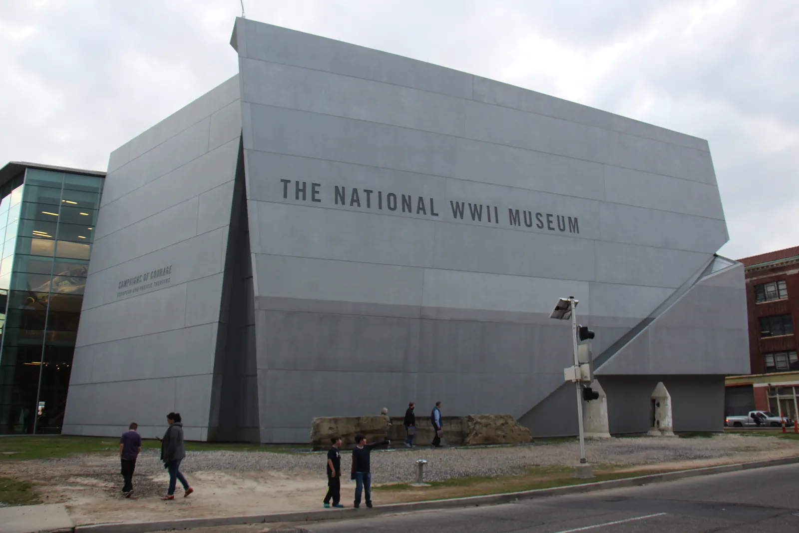 The angular grey stone facade of the National WWII Museum in New Orleans, its name cut into the wall above the street, with people walking past on the pavement