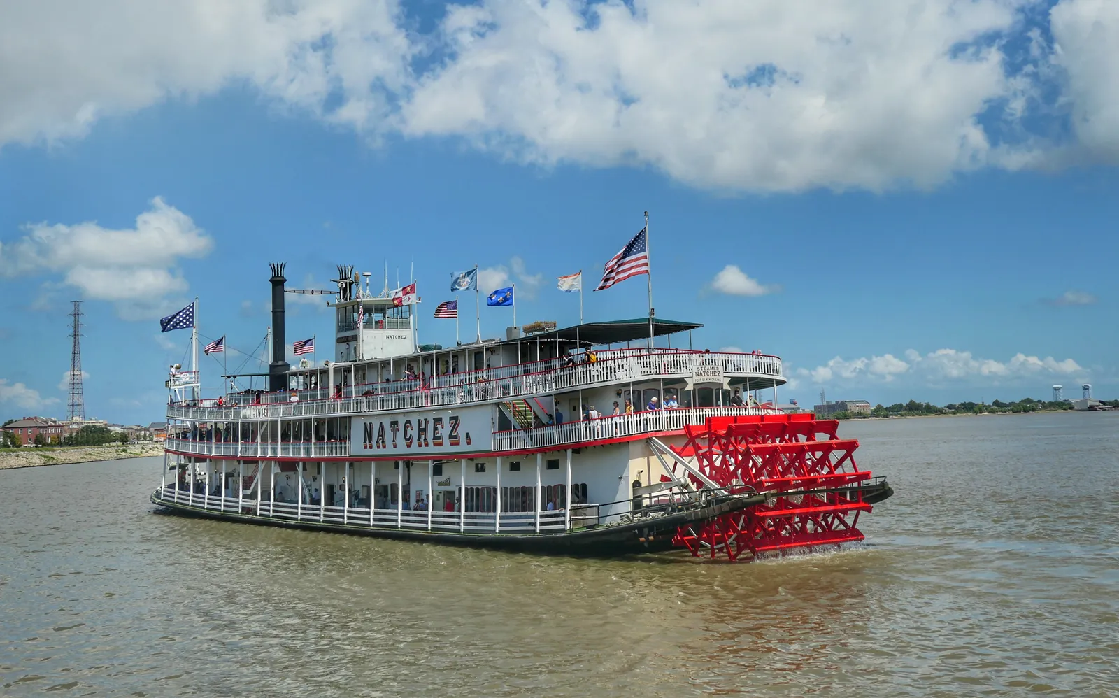 The Steamboat Natchez on the Mississippi at New Orleans, a white sternwheeler with two black funnels, flags along the upper deck and a bright red paddlewheel at the stern