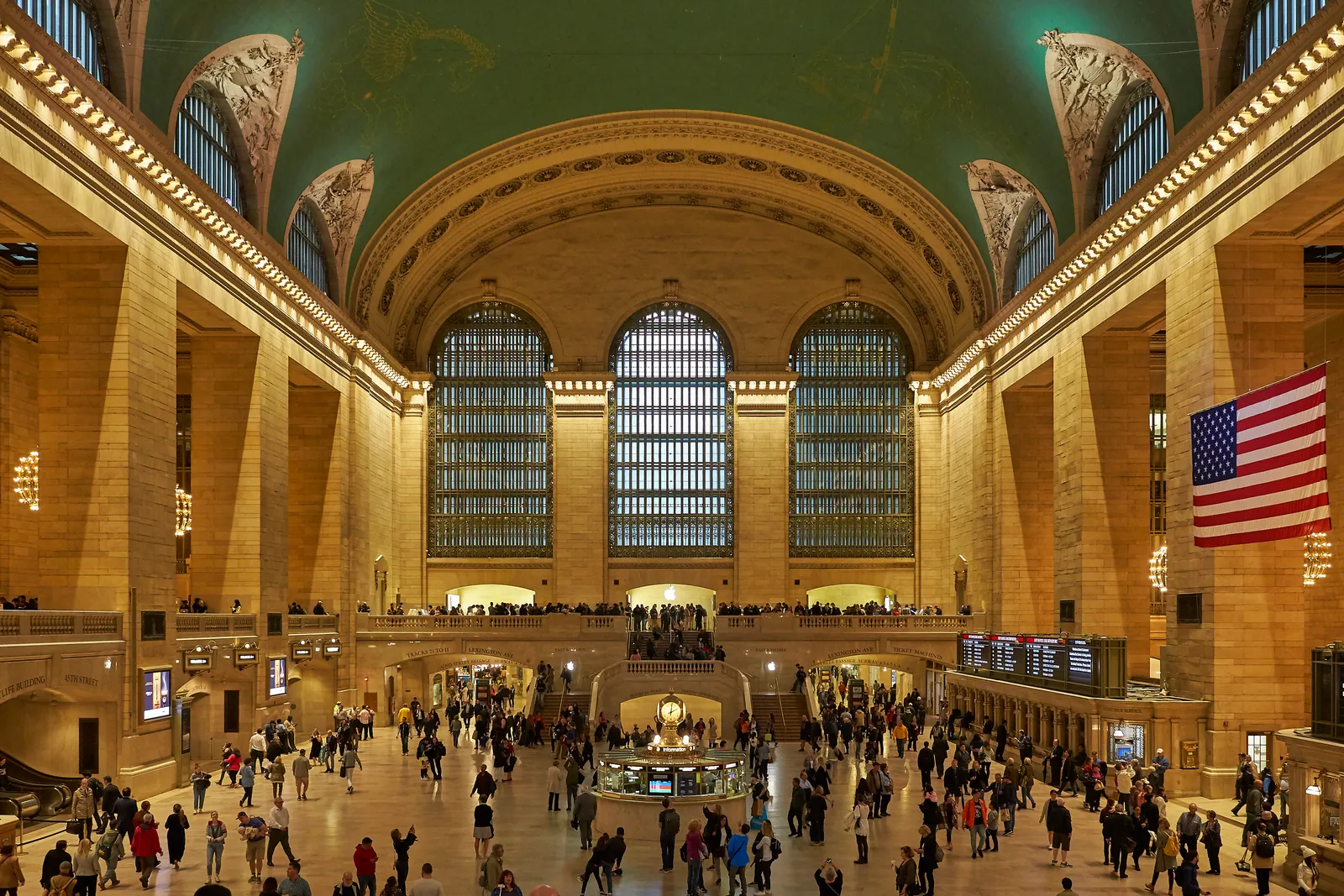 The Main Concourse of Grand Central Terminal, its green painted ceiling above three tall arched windows, with the information booth and crowds of people on the marble floor below