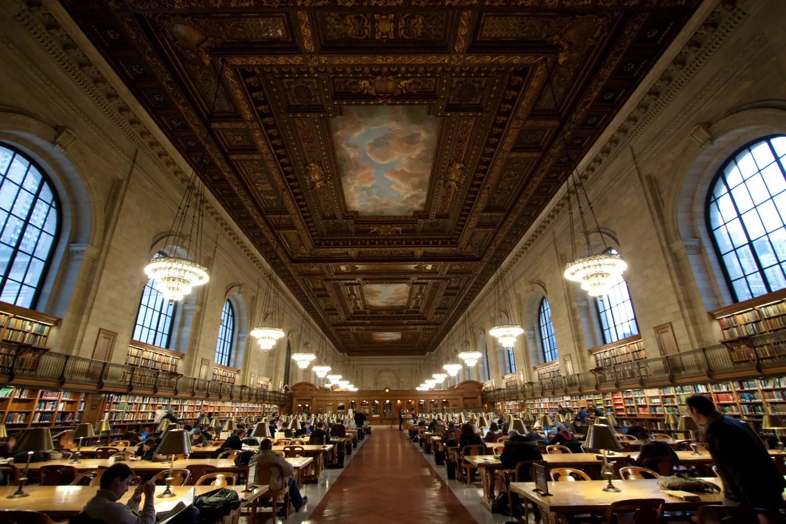 The Rose Main Reading Room at the New York Public Library, two long rows of oak tables with brass lamps under a painted cloud ceiling and hanging chandeliers