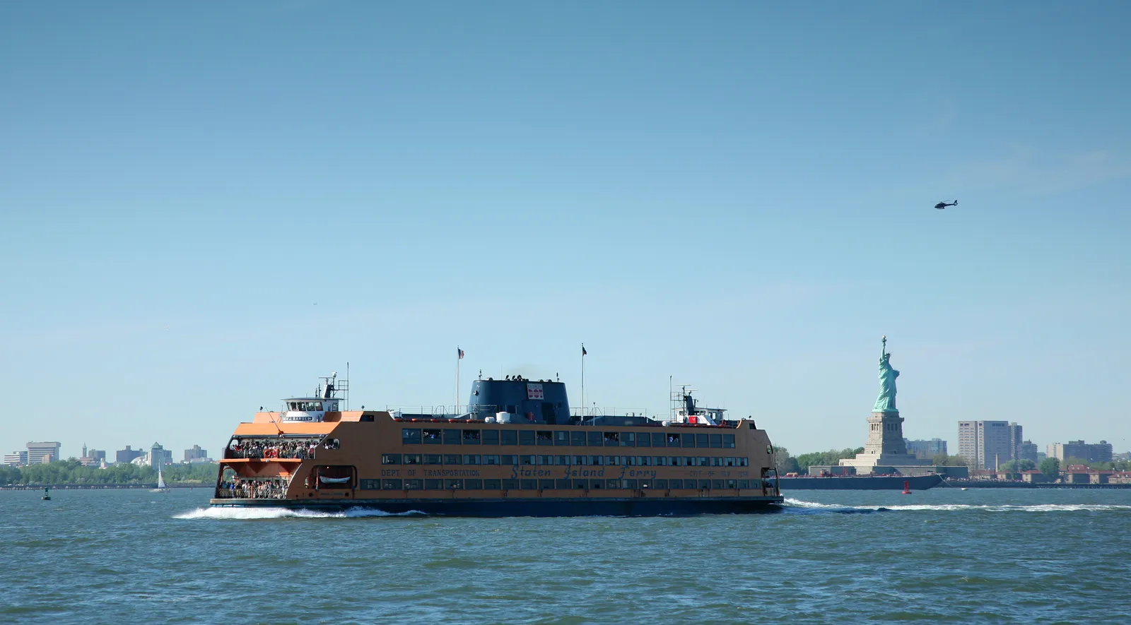 An orange Staten Island Ferry crossing New York Harbor in daylight, with the Statue of Liberty on its pedestal visible in the water behind it