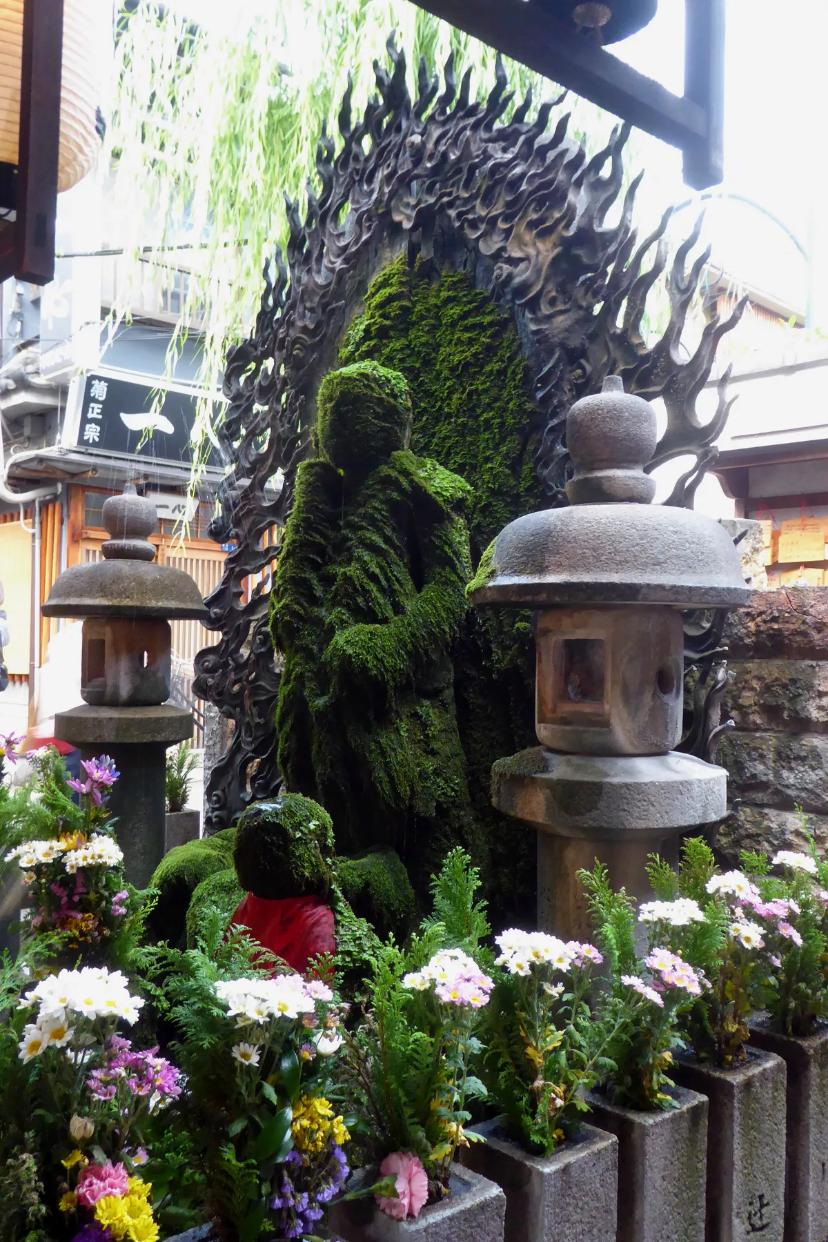 The standing Fudo Myo-o figure at Hozen-ji in Osaka, completely covered in thick green moss so that only the outline of the face and robes is readable, framed by a carved flame halo, with stone lanterns and fresh flowers in front of it