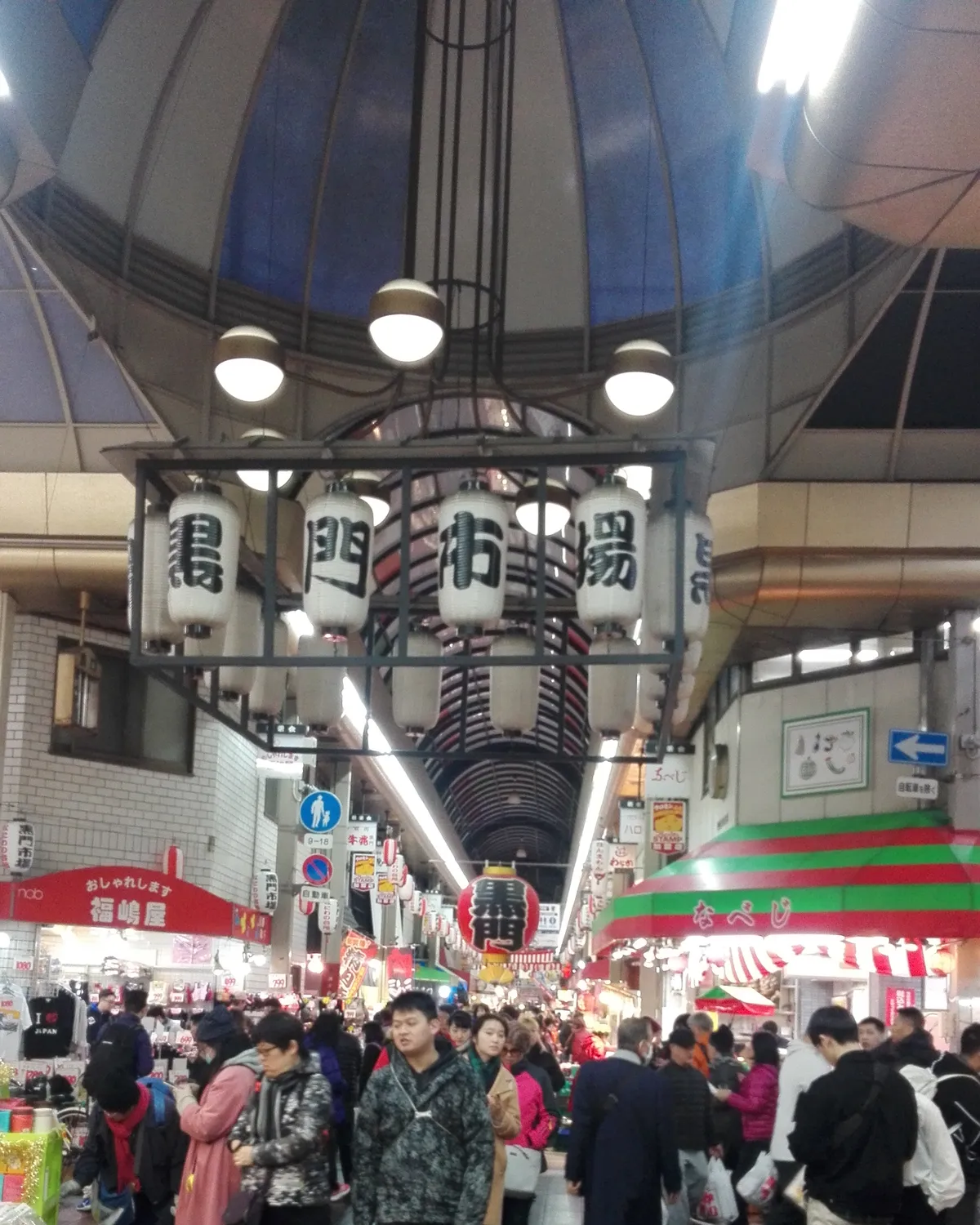 The Kuromon Ichiba arcade in Osaka, a covered shopping street packed with people under a long vaulted roof, with a cluster of white paper lanterns hanging from a frame in the foreground and stall awnings running away down both sides