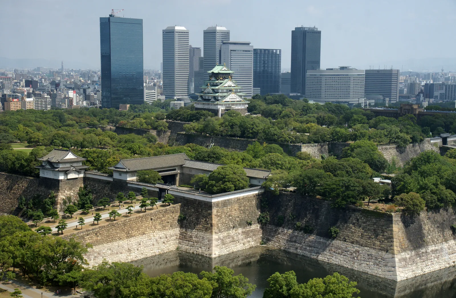 Osaka Castle seen from above, the white and green keep standing among trees inside a ring of sloped stone walls and a water-filled moat, with the office towers of Osaka Business Park rising directly behind it