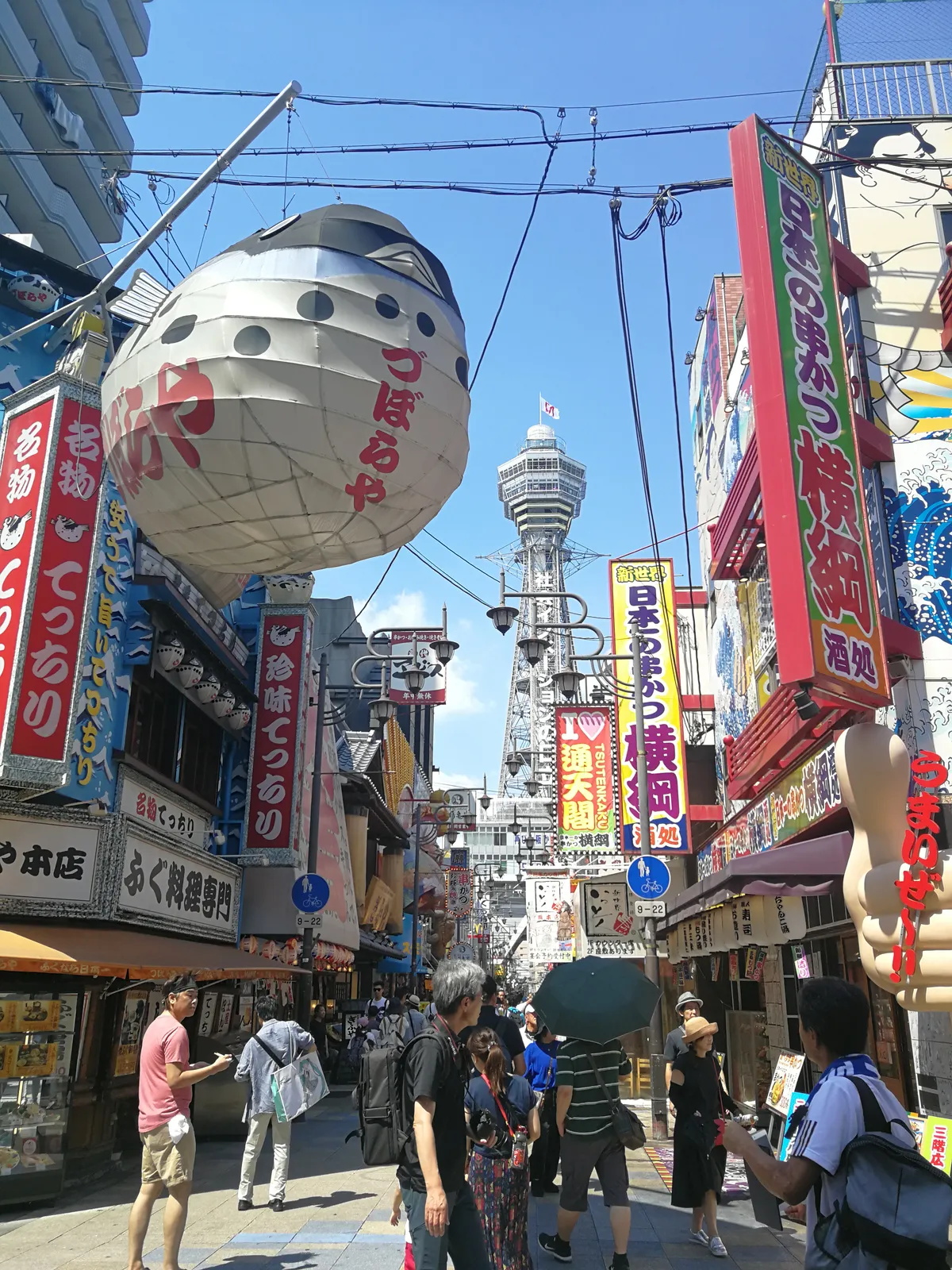 A street in the Shinsekai district of Osaka crowded with painted restaurant signboards and a large pufferfish lantern hanging overhead, with the steel lattice of Tsutenkaku tower rising at the end of the street against a blue sky