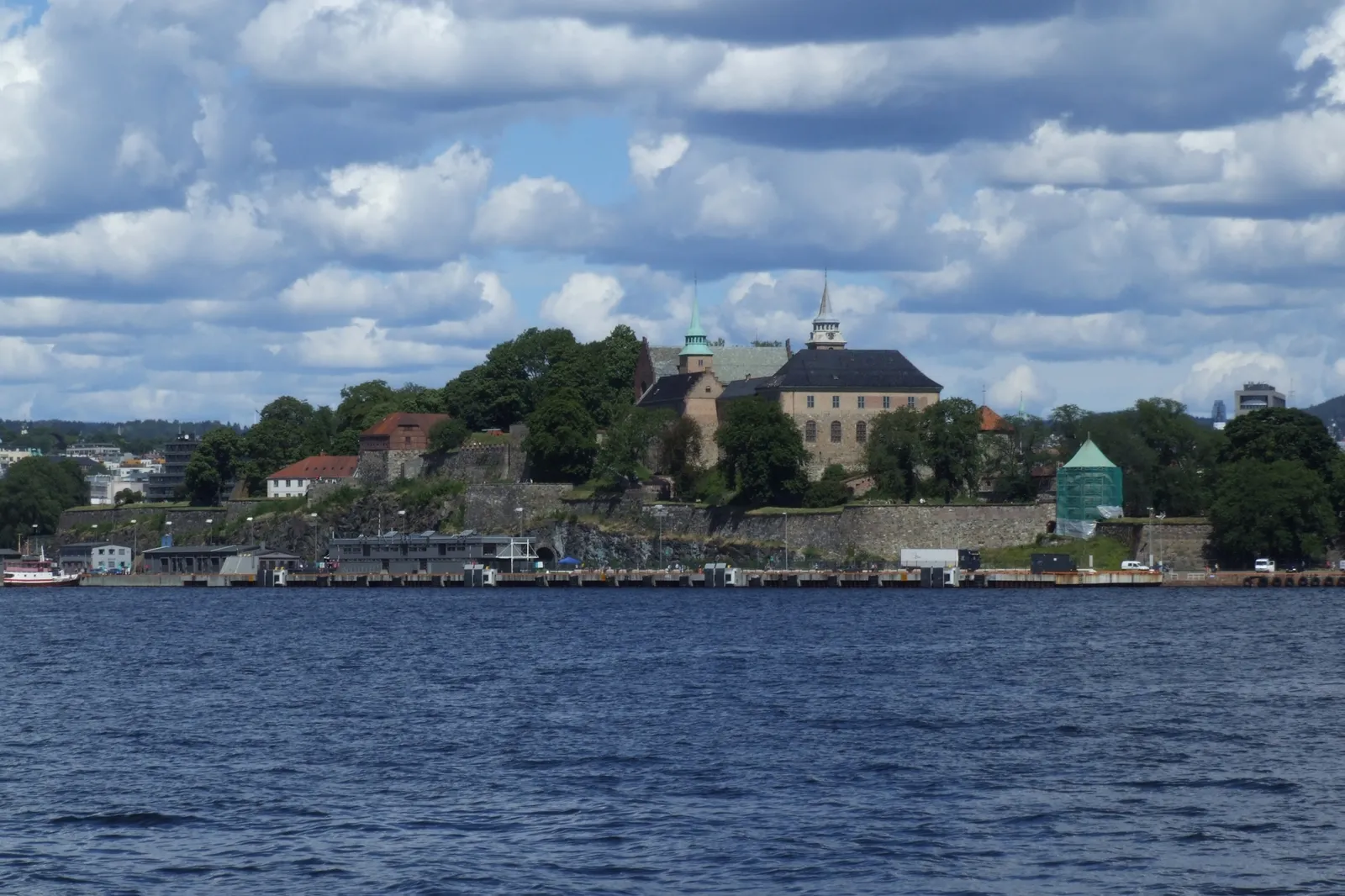 Akershus Fortress in Oslo seen across the water, the castle's green copper spire and dark roofs standing among trees on a low headland above stone ramparts and the harbour quay