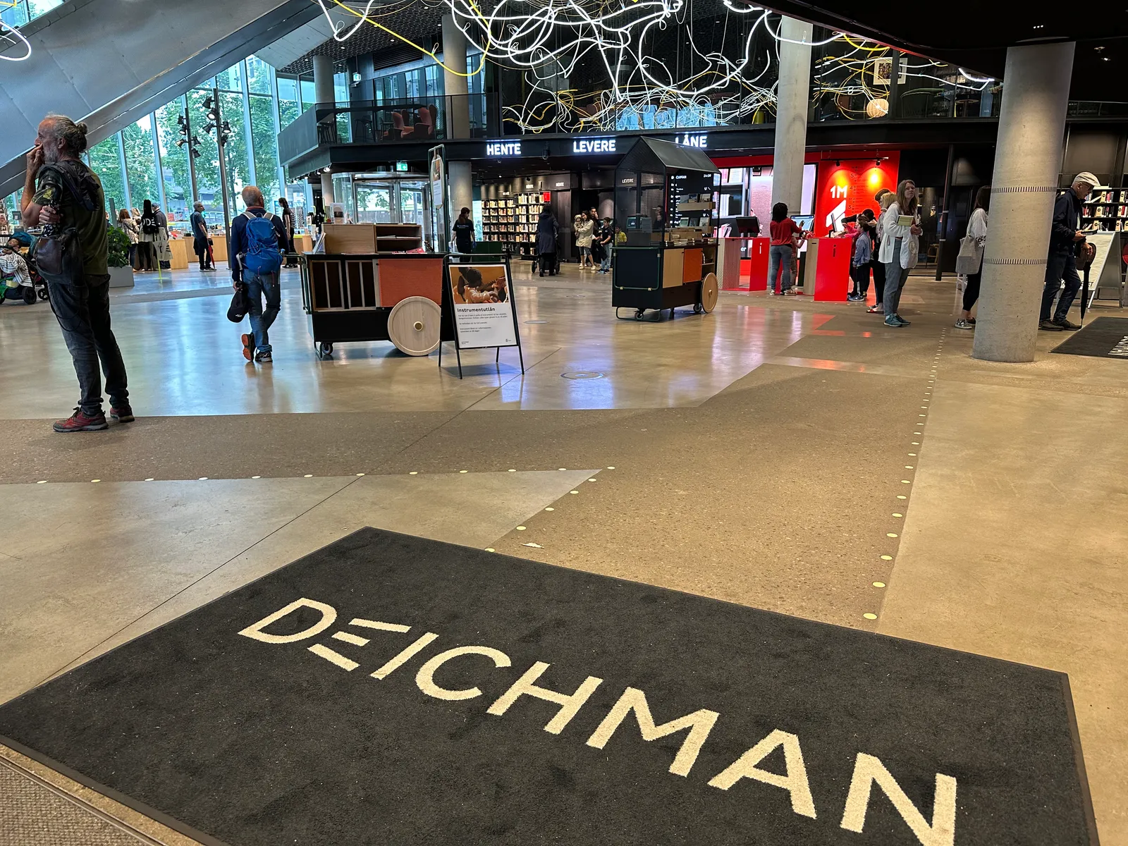 The ground floor of the Deichman Bjørvika library in Oslo, a wide polished concourse with the library's name on a mat in the foreground, shelves and a service desk beyond, and people walking through