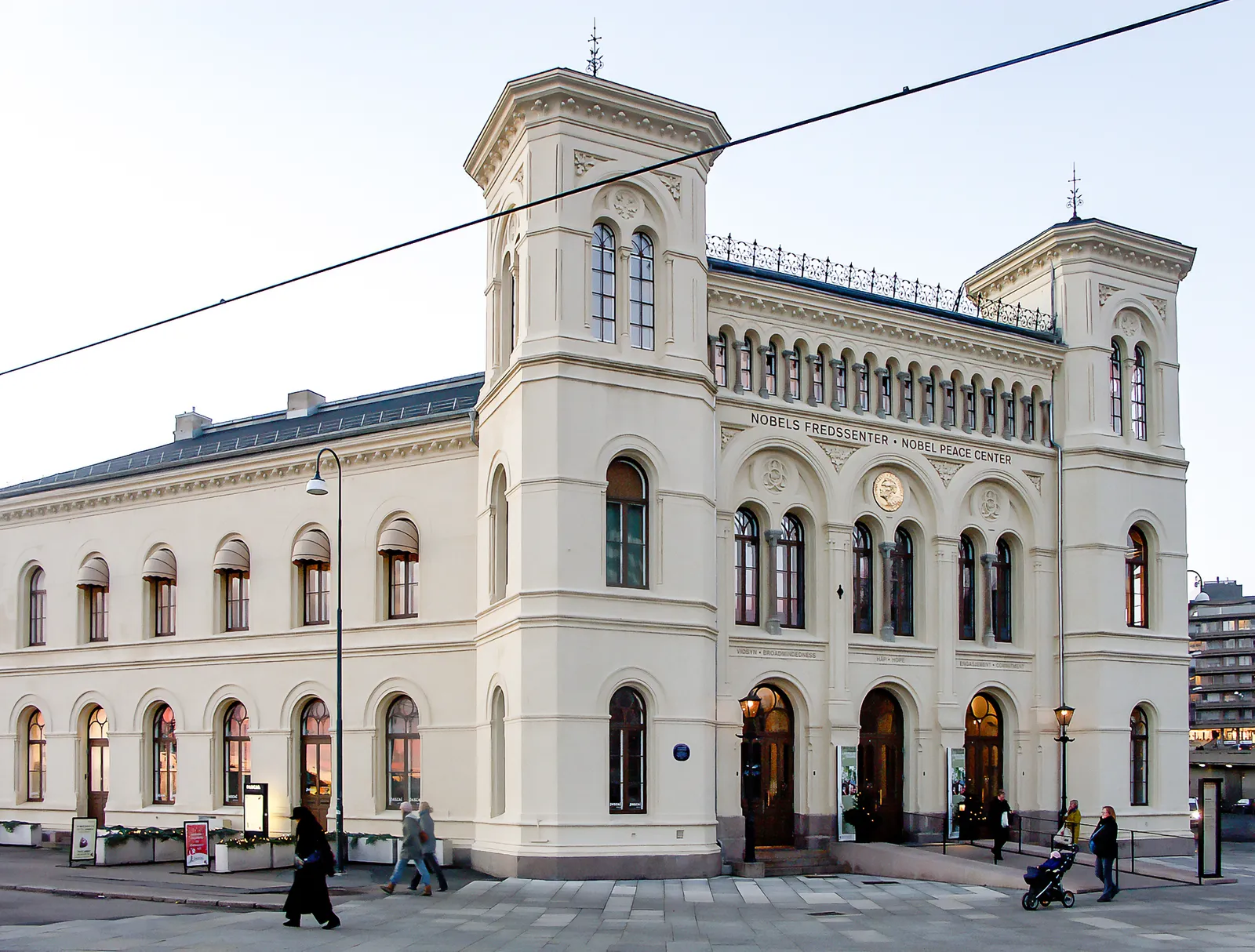 The Nobel Peace Center in Oslo, a pale yellow 19th-century railway station building with two corner towers and arched windows, its name lettered across the front above the entrance arches