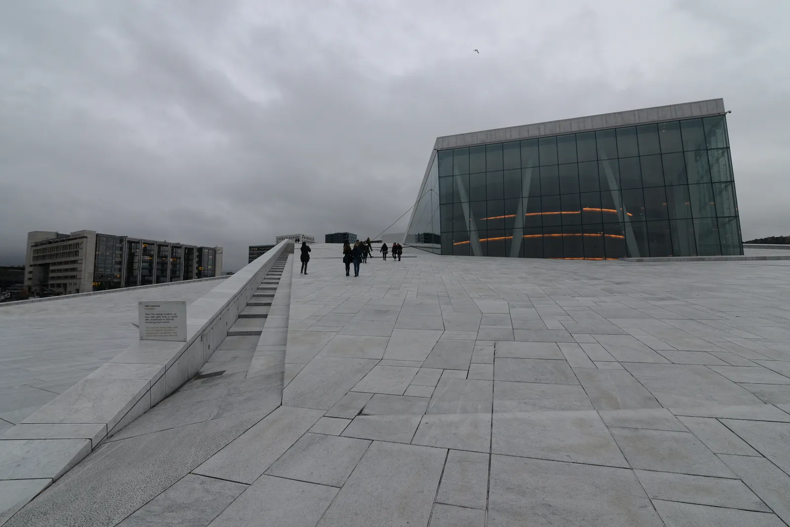 The marble roof of the Oslo Opera House from ground level, a wide pale stone ramp climbing towards the angular glass tower of the stage house, with people walking up it