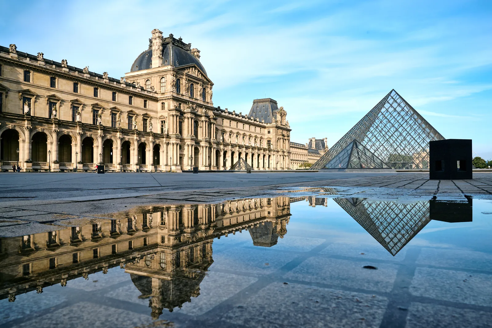 The glass pyramid in the Cour Napoléon of the Louvre in Paris, with the carved stone facade of the Denon wing running along one side and rainwater on the paving reflecting both