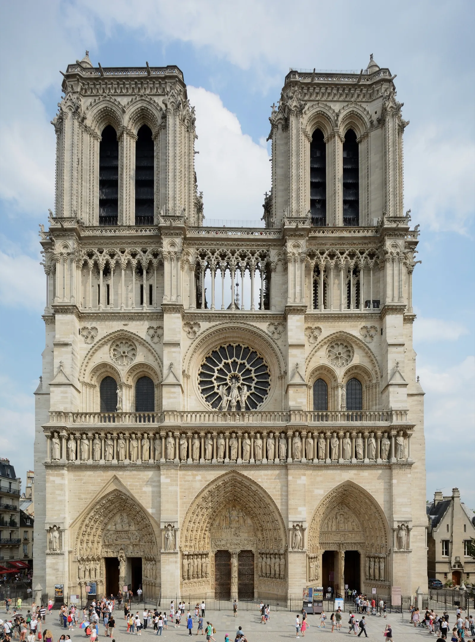 The west front of Notre-Dame de Paris, its two square towers above the rose window and the row of stone kings, with visitors crossing the parvis at the foot of the three portals