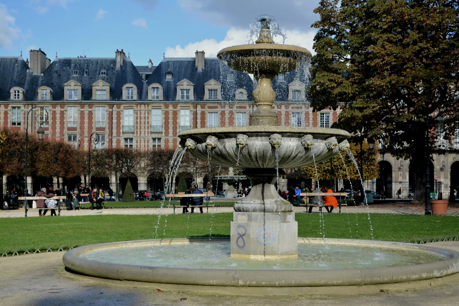 A stone fountain playing in the garden of the Place des Vosges in Paris, with the square's red brick and cream stone pavilions and steep slate roofs behind it and people sitting on the grass