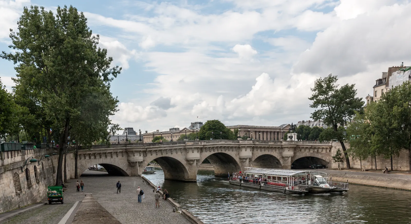 The Pont Neuf in Paris, its stone arches spanning the Seine, with a riverboat passing beneath and people walking the paved quay along the water's edge