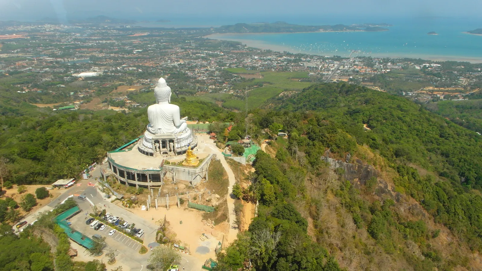 The white marble Buddha seated on the ridge of Nakkerd Hill in Phuket seen from above, with forested slopes falling away on both sides and Chalong Bay and the island's southern coast behind it