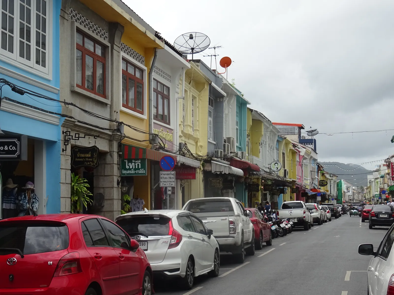 Thalang Road in Phuket Town, a row of two-storey shophouses painted yellow, blue and cream with shuttered upper windows and shop awnings over the arcade, cars parked along the kerb