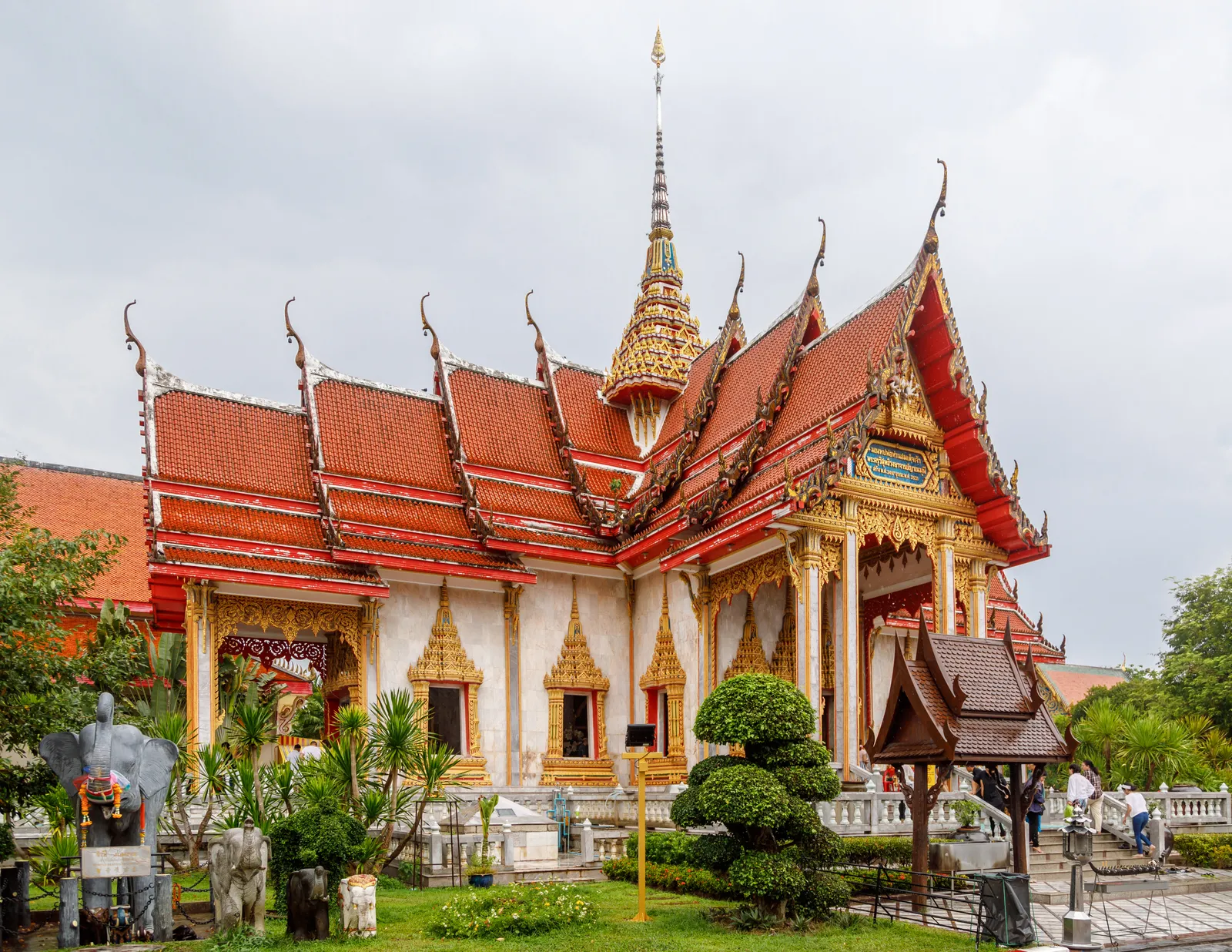 The main hall of Wat Chalong in Phuket, tiered orange and red roofs edged in gold with a gilded spire above them, standing over a lawn with clipped shrubs and a small pavilion
