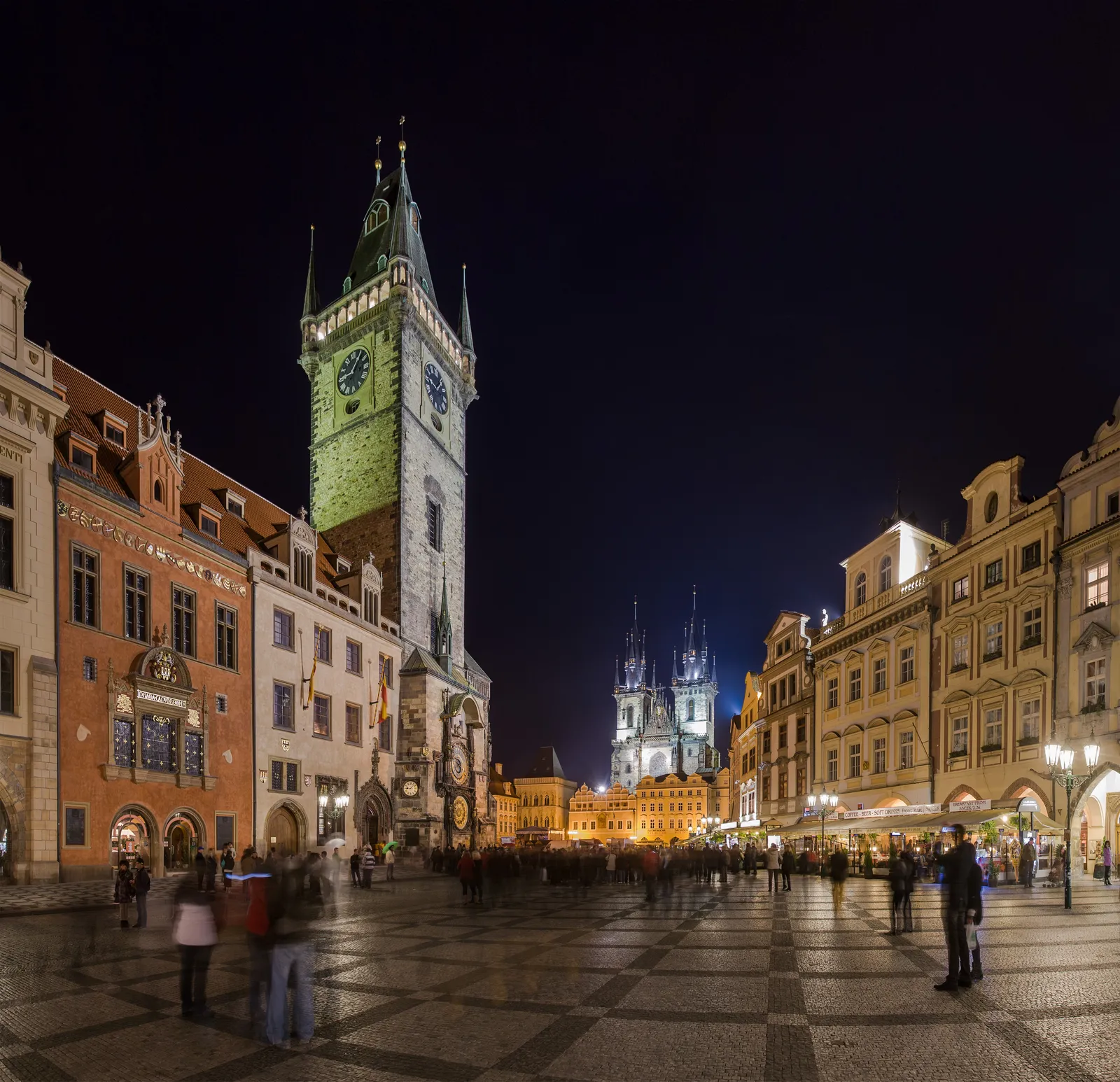 Old Town Square in Prague after dark, with the lit Gothic tower of the Old Town Hall on the left, the twin spires of the Týn Church behind the pastel houses opposite, and people crossing the paved square