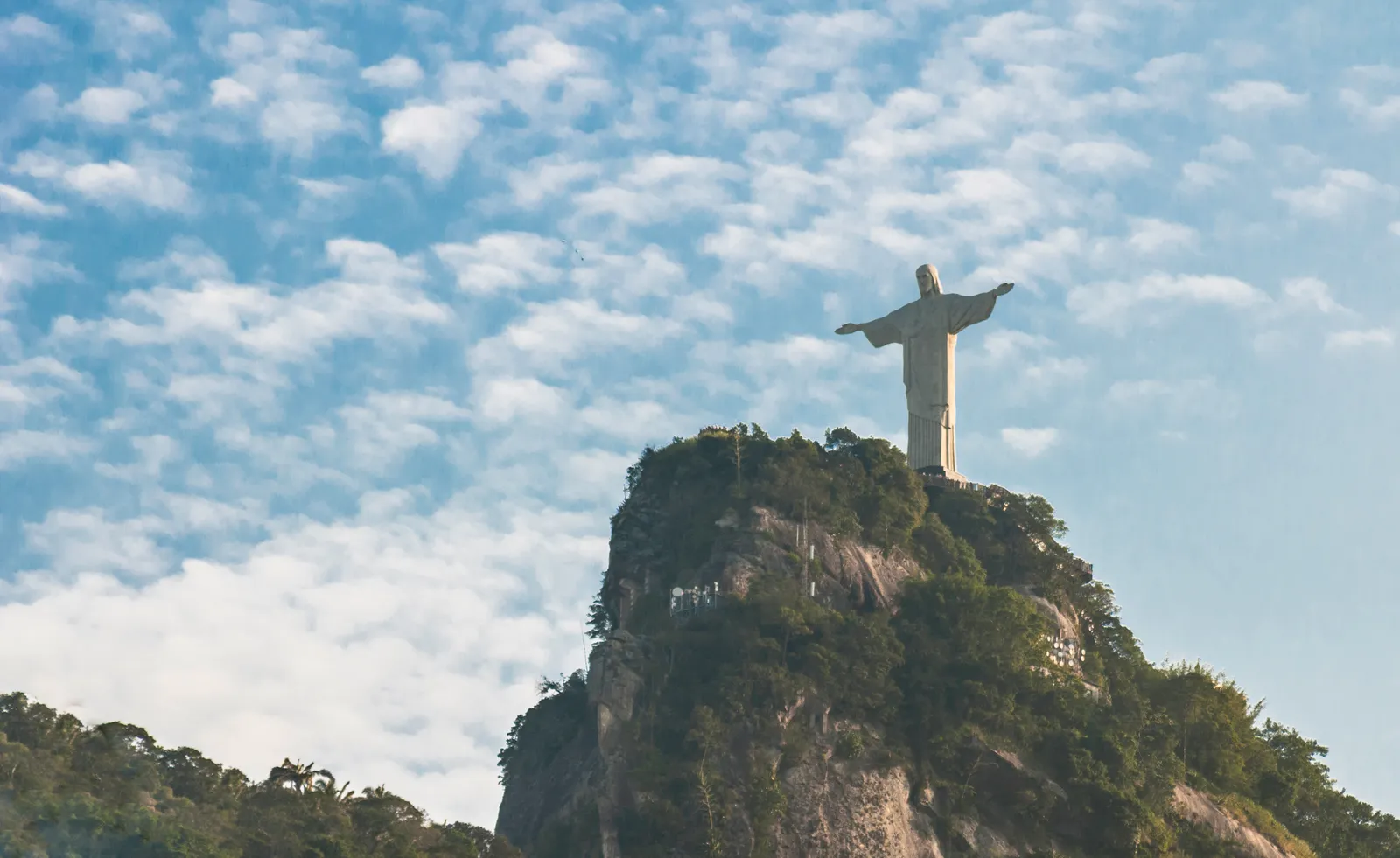 The Christ the Redeemer statue standing with arms outstretched on the bare rock summit of Corcovado in Rio de Janeiro, forest covering the slopes below it and broken cloud behind