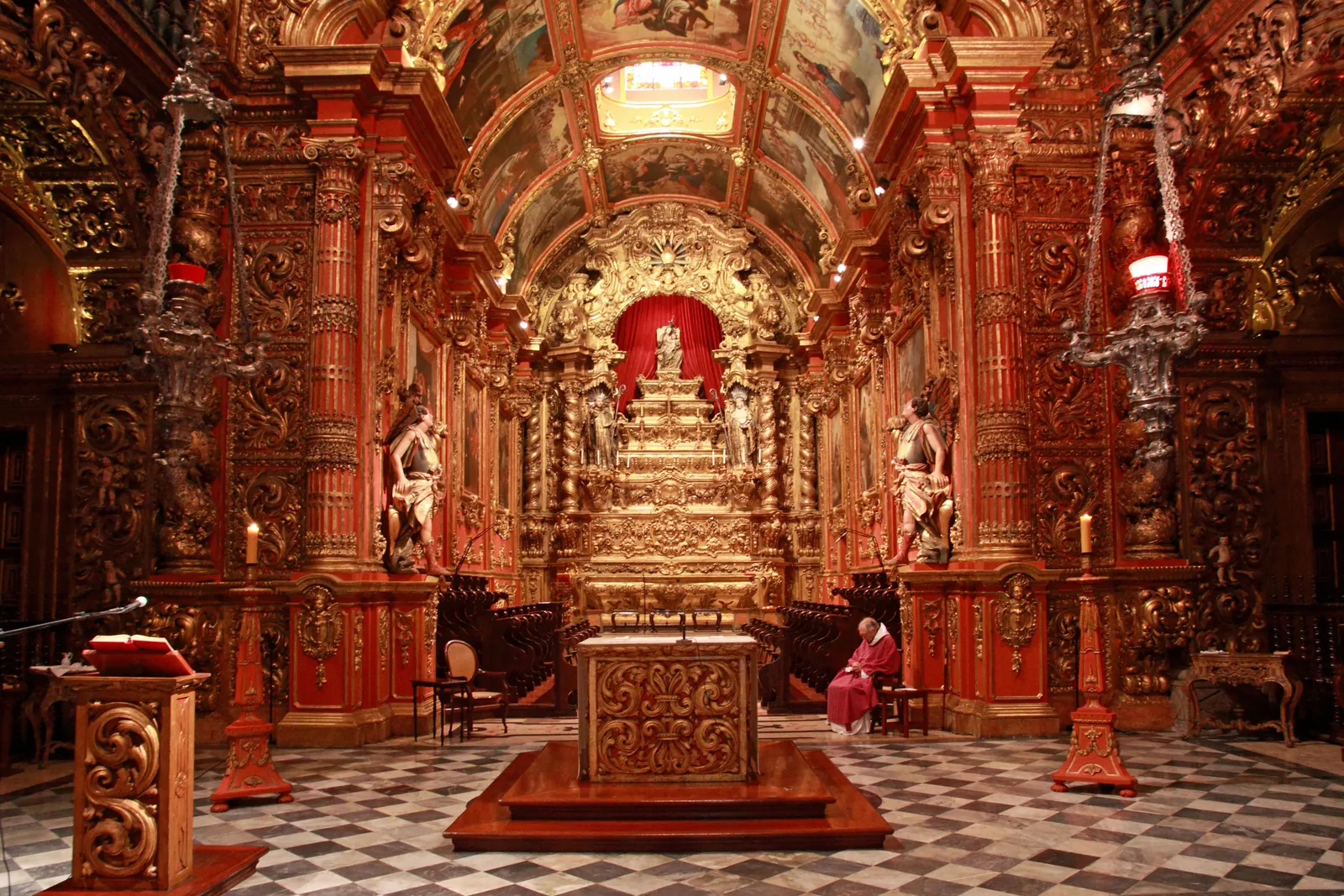 The interior of the São Bento monastery church in Rio de Janeiro, its walls, columns and altar screen carved and covered in gold leaf, with a painted ceiling above and a black and white tiled floor below