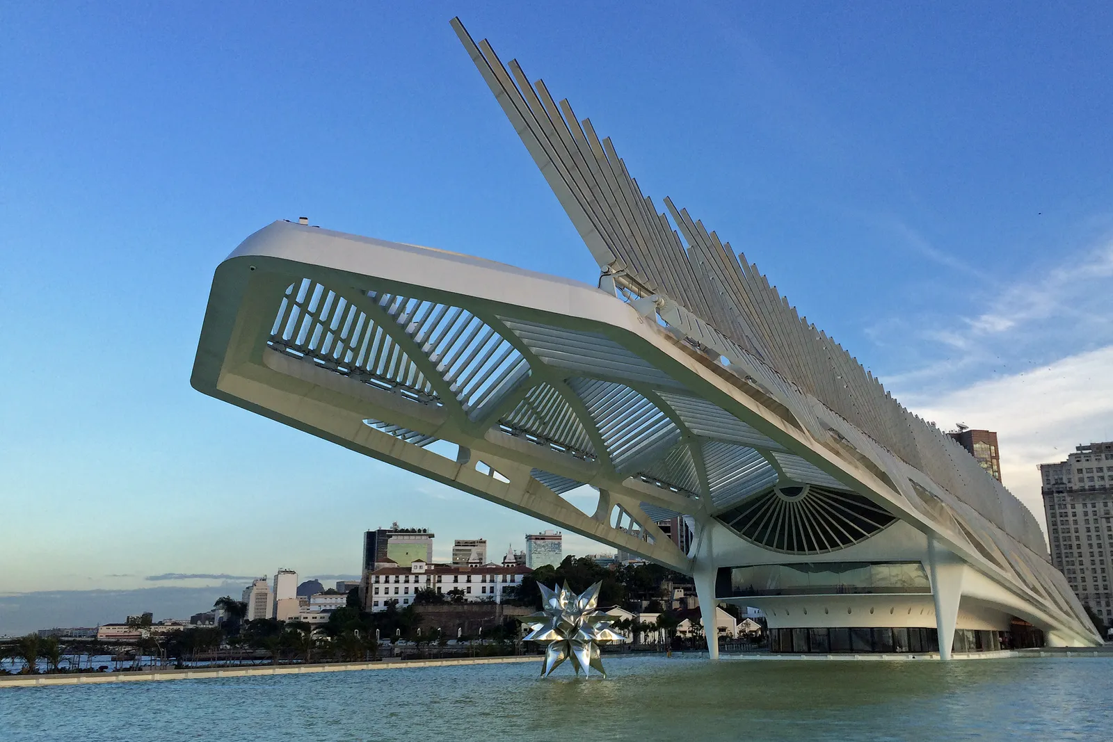 The Museum of Tomorrow on the Rio de Janeiro waterfront, a long white building whose roof cantilevers out over a reflecting pool with rows of angled fins along the top of it