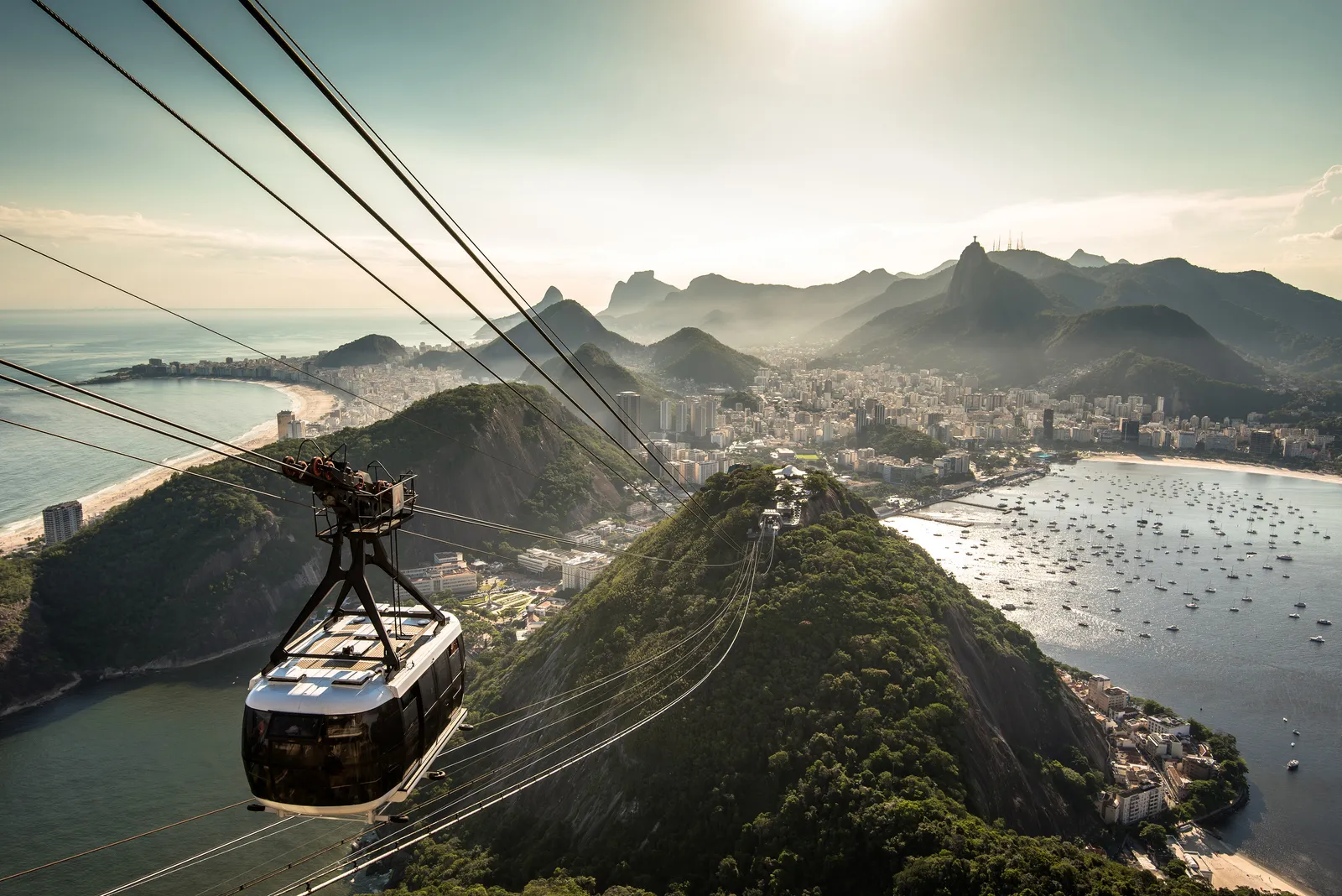 A glass-sided cable car cabin climbing towards Sugarloaf Mountain in Rio de Janeiro, with the forested dome of Morro da Urca below it, Botafogo bay full of moored boats to the right and the mountains behind the city in the haze