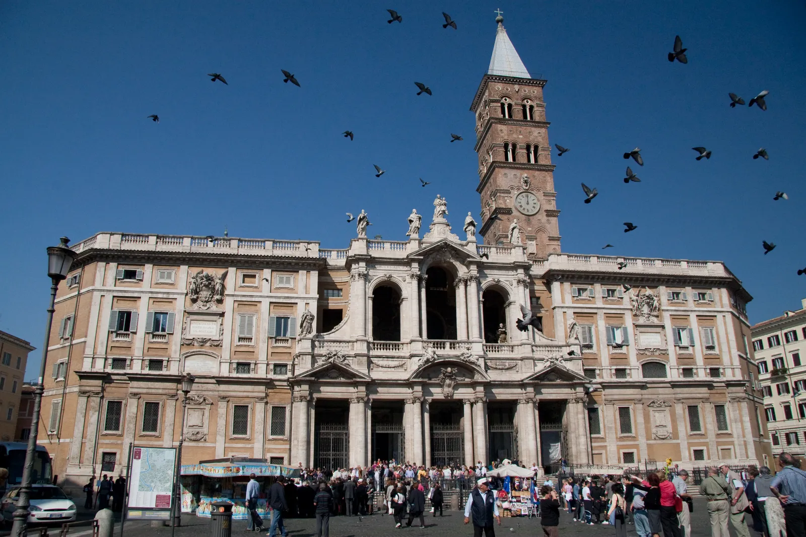 The front of the Basilica of Santa Maria Maggiore in Rome, its pale stone loggia and columns under a blue sky, with the brick medieval bell tower rising behind and pigeons crossing the square