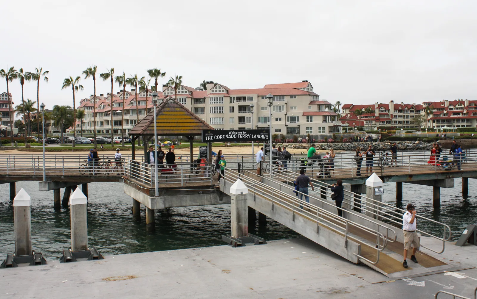The pier and covered shelter at the Coronado Ferry Landing, with palms and red-roofed buildings along the shore behind it