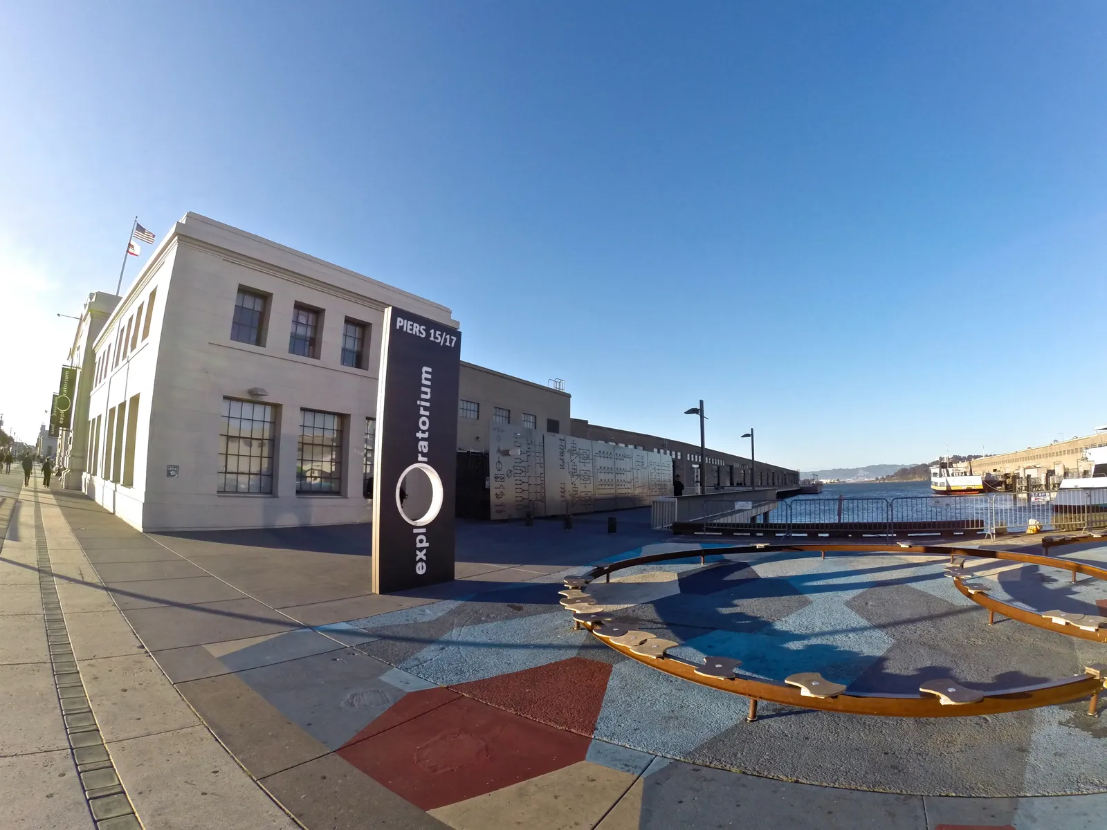 The Exploratorium on Pier 15 in San Francisco, a long low white pier shed with the museum's name on a black sign board and a coloured exhibit installed on the open plaza beside it