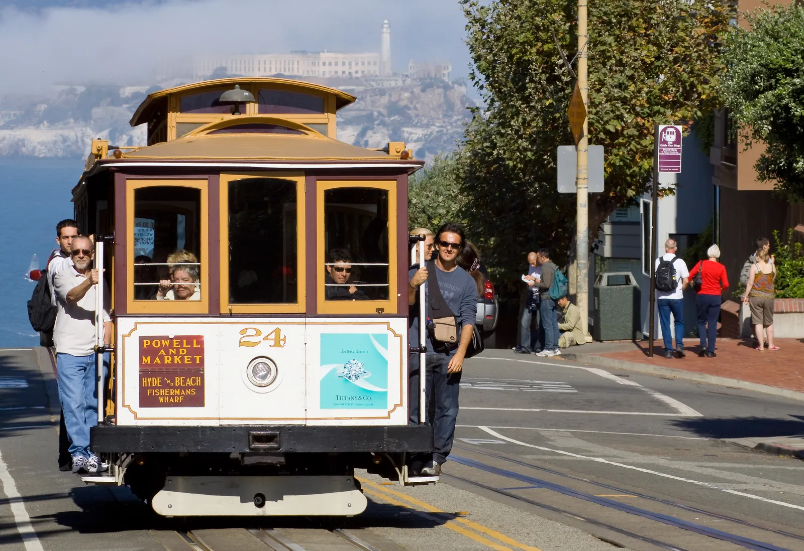 A Powell-Hyde cable car climbing Hyde Street in San Francisco with passengers standing on the running boards, the bay and Alcatraz Island visible in the haze behind it
