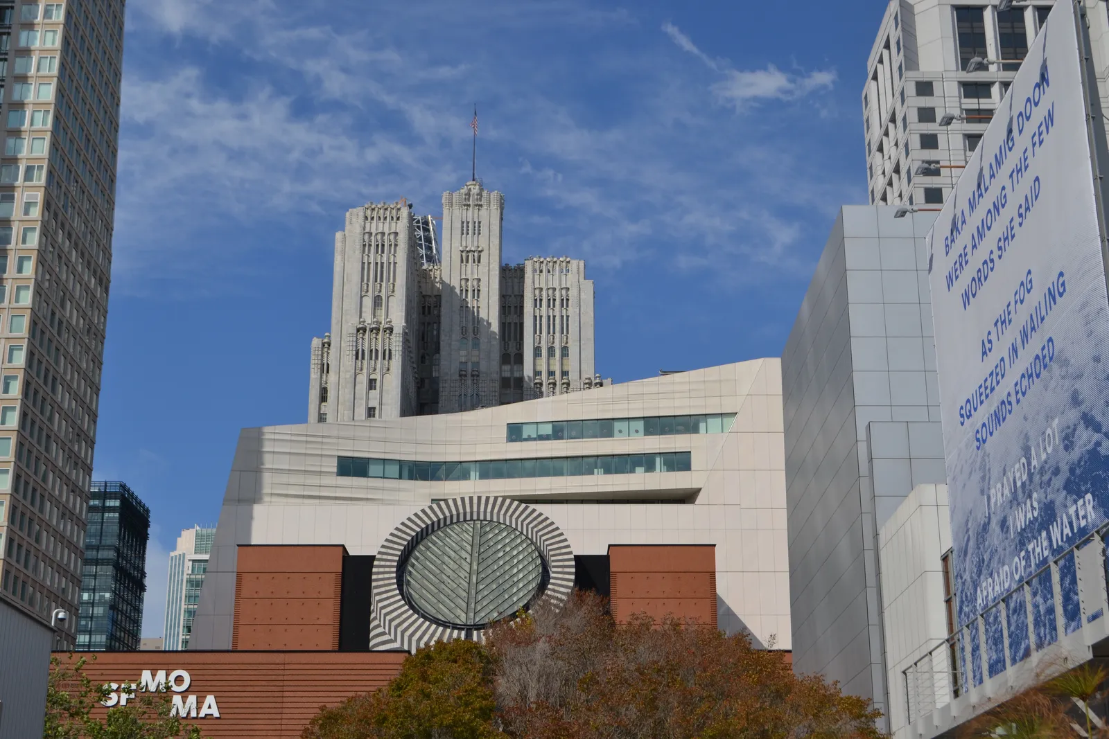 The San Francisco Museum of Modern Art from Third Street, its red brick block with a black and white striped oculus in front of the taller white addition behind, under a blue sky
