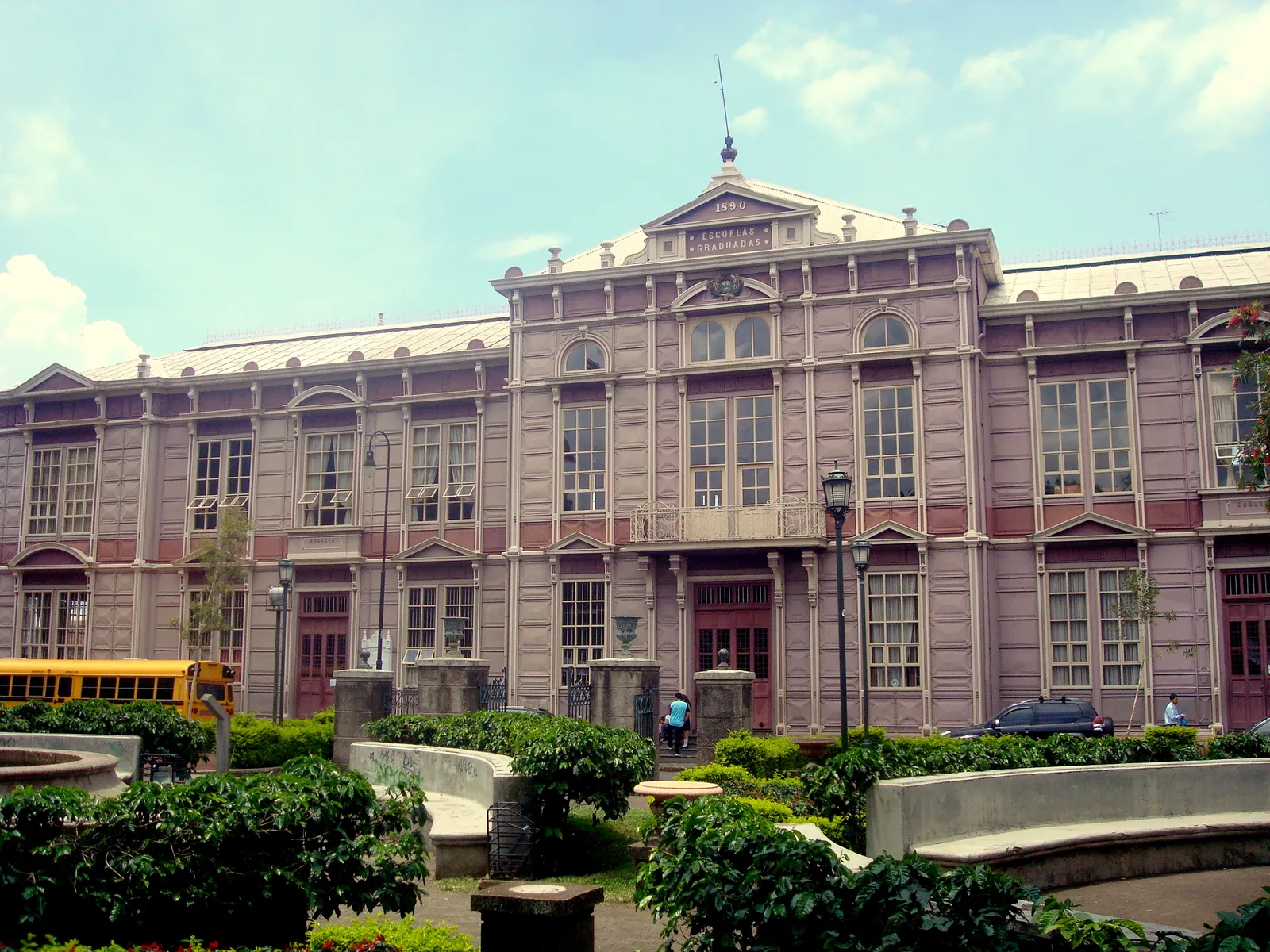 The long wrought-iron facade of the Metallic Building in San Jose, a two-storey prefabricated school painted dusty pink, with a yellow school bus parked at one end and gardens in front