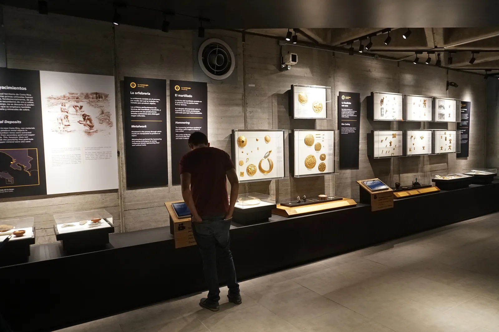 A visitor looking into lit display cases of pre-Columbian gold set against bare concrete walls in the underground gold museum in San Jose