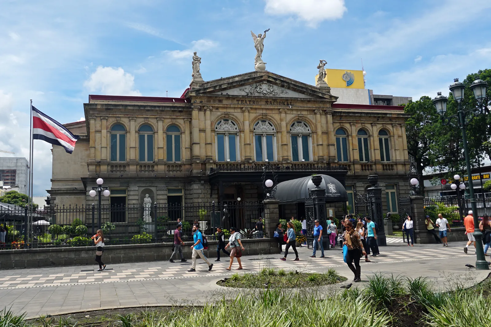 The stone facade of San Jose's National Theatre with statues along its roofline, a Costa Rican flag flying at the left and people crossing the paved square in front of it