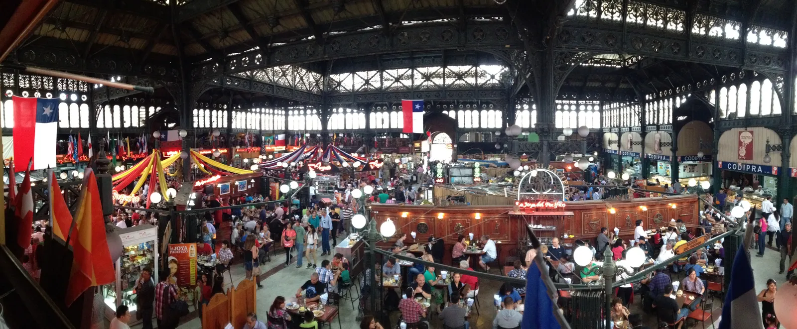 The interior of Santiago's central market hall, with its riveted iron roof trusses arching over a floor packed with restaurant tables and Chilean flags strung overhead