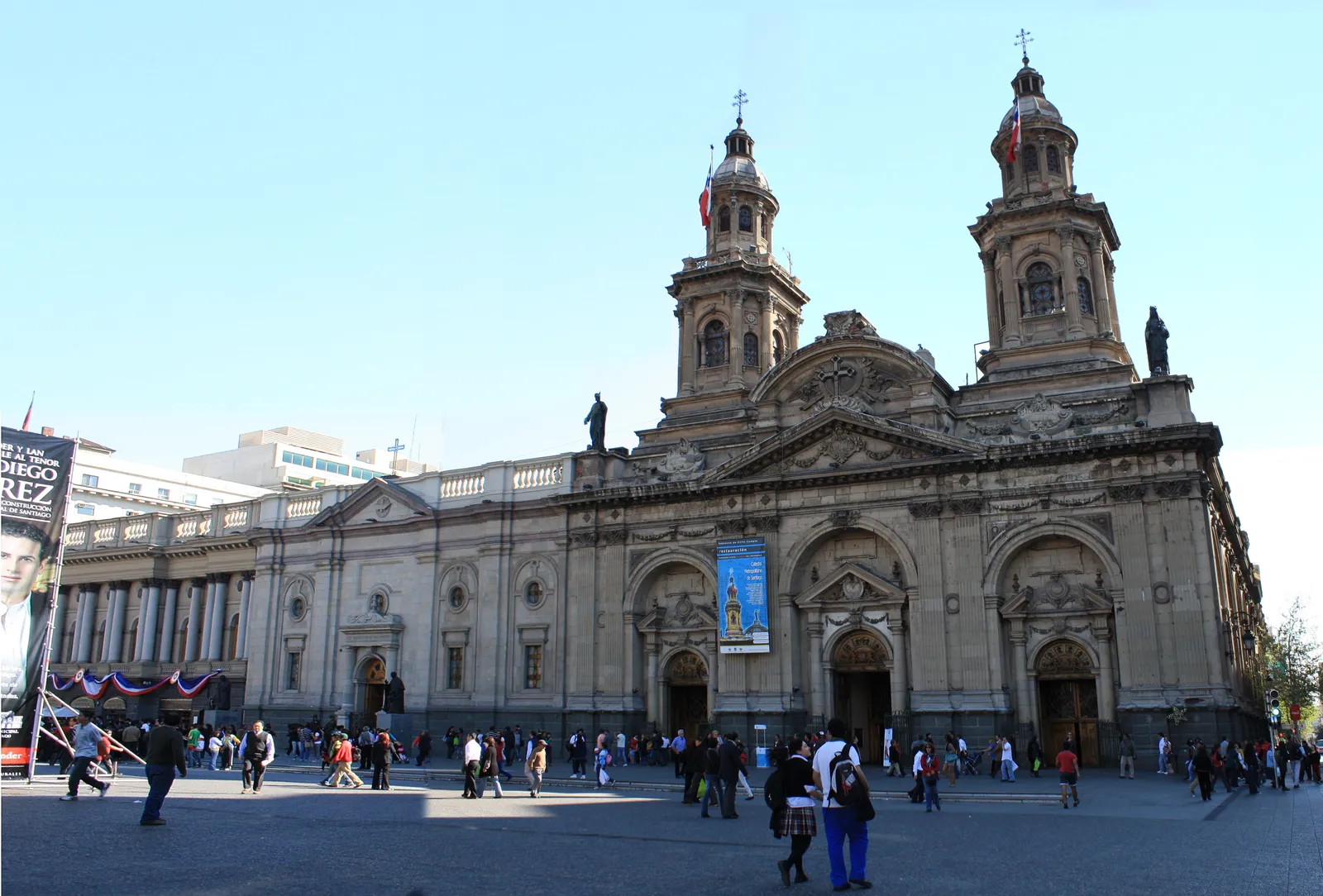 The neoclassical facade and twin bell towers of Santiago's Metropolitan Cathedral seen across the paved Plaza de Armas, with people walking in front of it