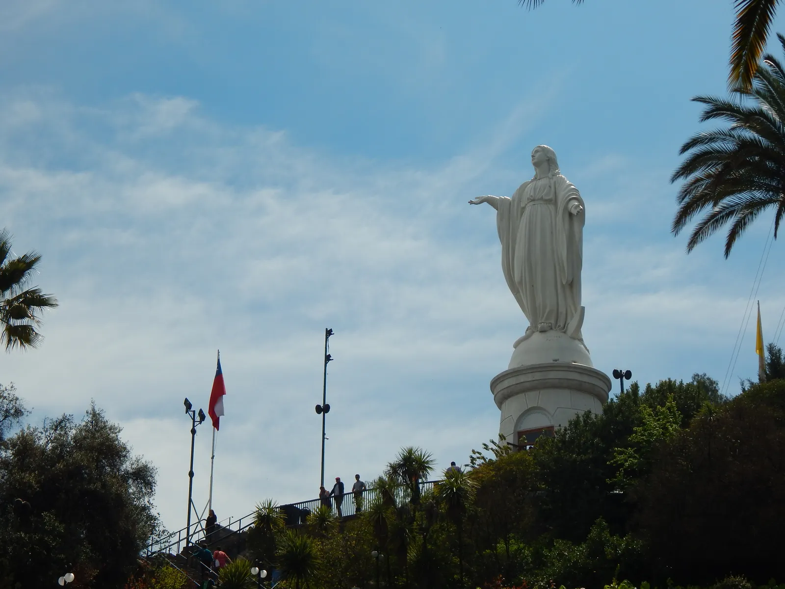 The white statue of the Immaculate Conception on its round pedestal at the top of San Cristóbal hill in Santiago, with palms, a Chilean flag and a railed viewing terrace below it