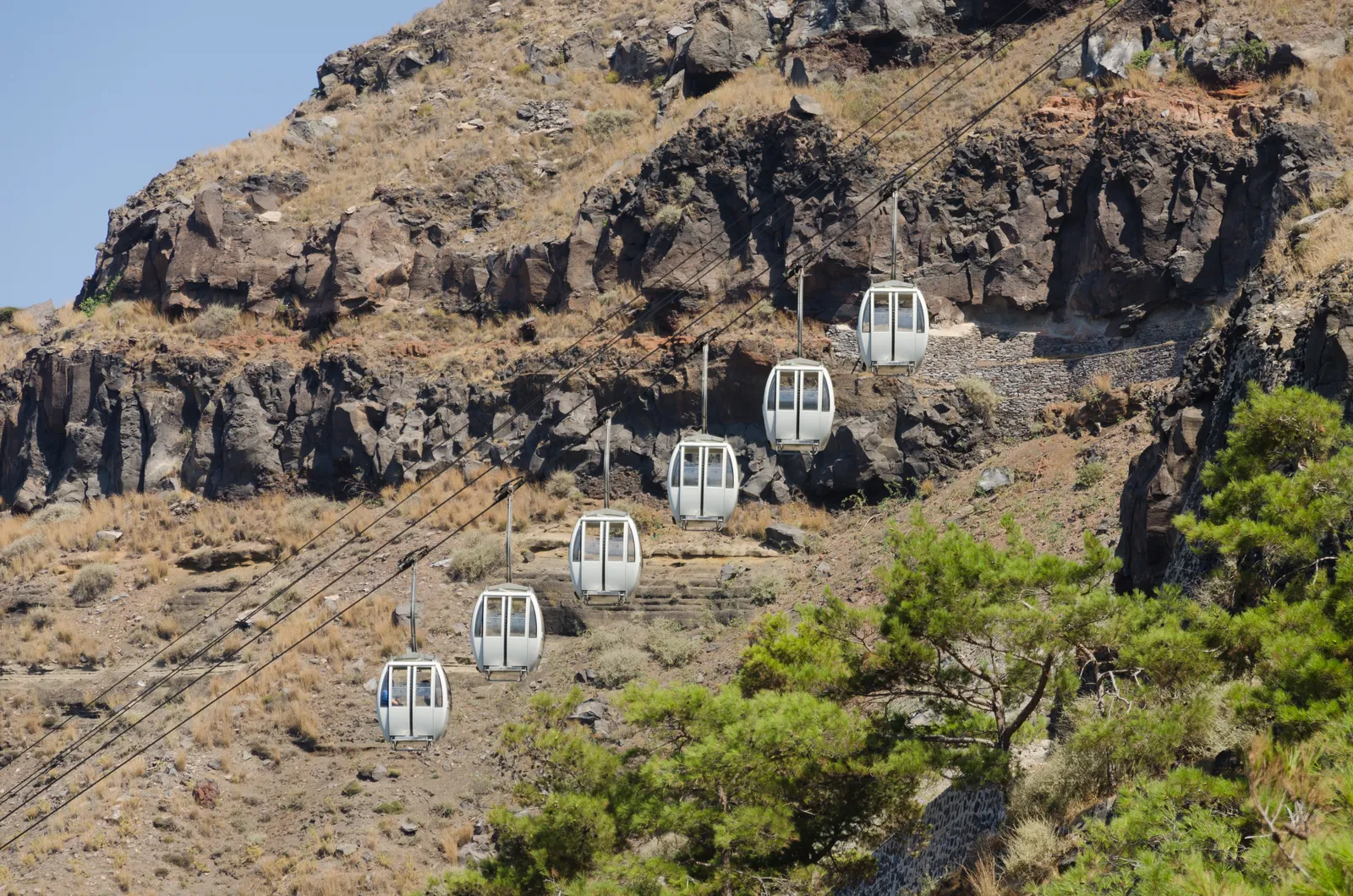 Six white cable car cabins strung down the bare volcanic cliff face between Fira and Santorini's Old Port, with the track cut into the rock beside them