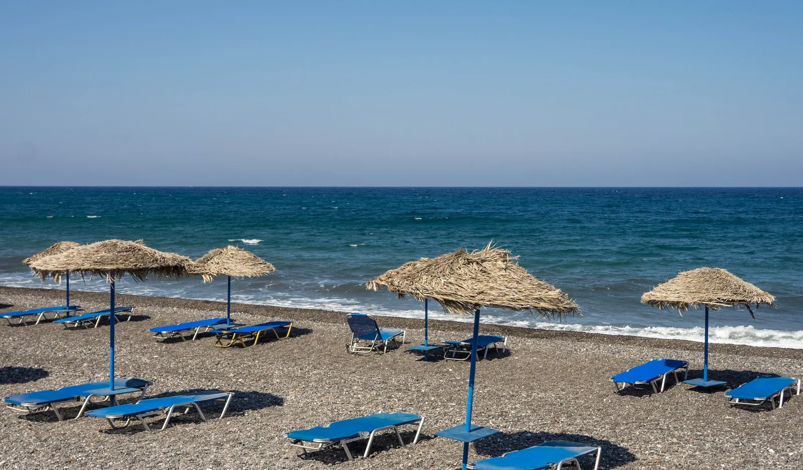 Kamari's dark grey volcanic sand with blue sunbeds under straw umbrellas and the open Aegean beyond
