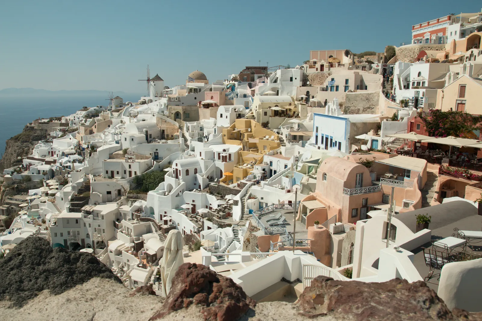 Oia seen from the castle at its western tip, white and pale pink houses stacked down the caldera slope with a stone windmill on the skyline
