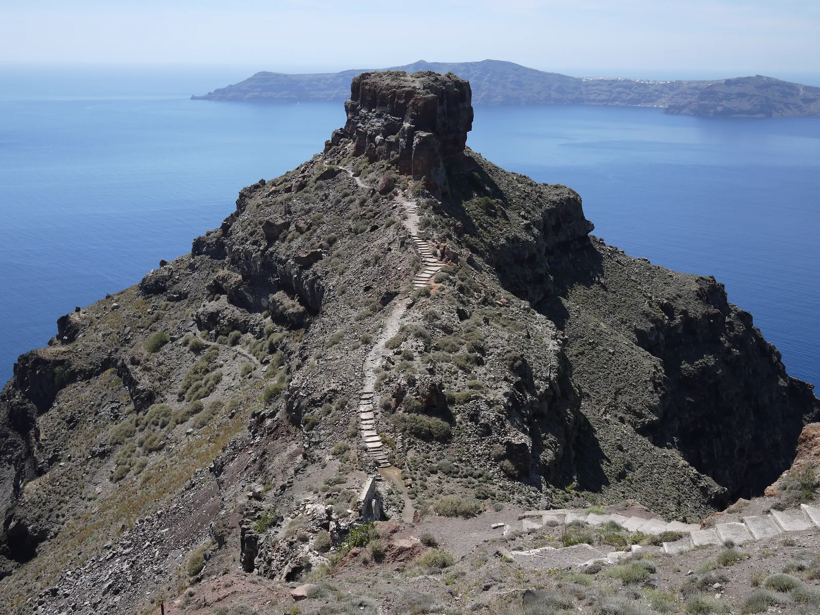 The Skaros headland jutting into Santorini's caldera, a bare brown rock with a stepped path zigzagging up its spine and the far crater wall across the blue water behind